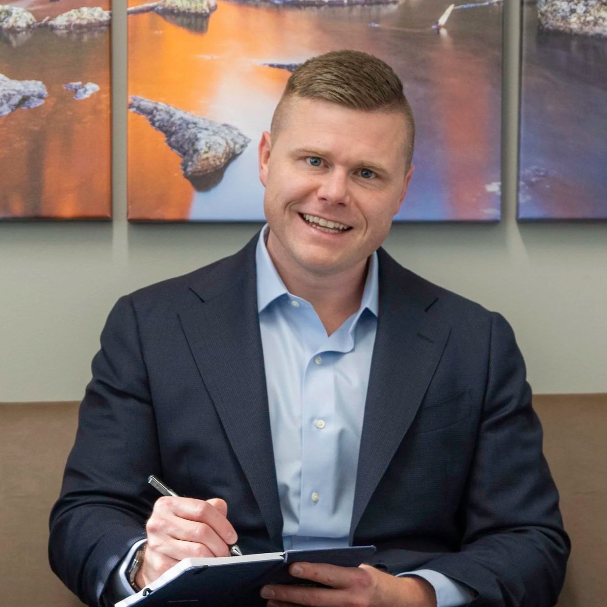 Jonathan Thomas in a dark suit and light blue shirt smiling while sitting at a table, holding a pen and a notebook, with a multi-panel landscape painting of rocks and water behind him.