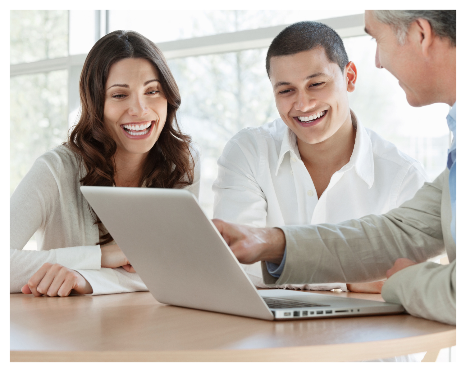 Three people, a woman and two men, sitting at a table looking at a laptop and smiling, in a bright office with large windows.