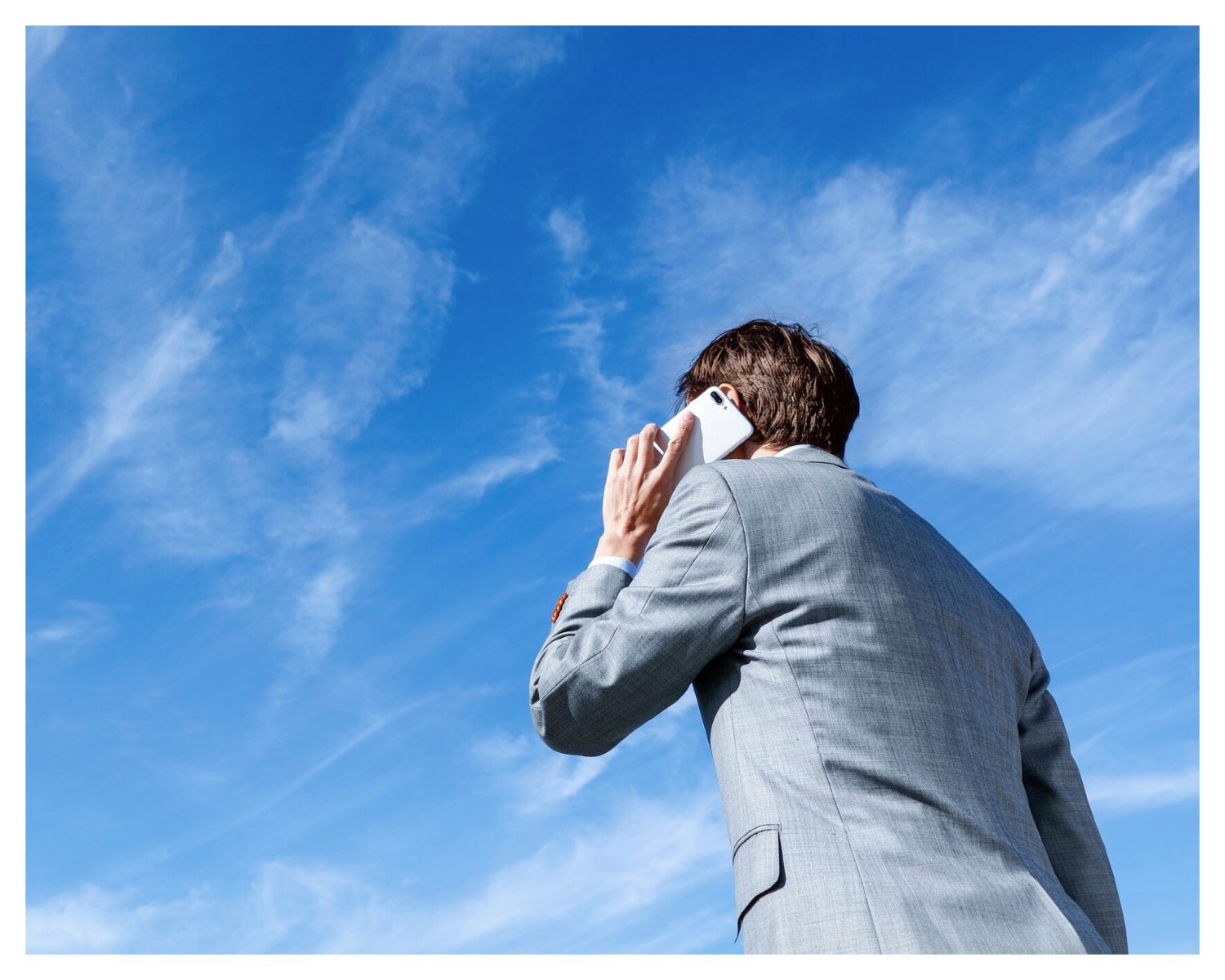 A man in a gray suit talking on a white cellphone against a blue sky with wispy clouds.