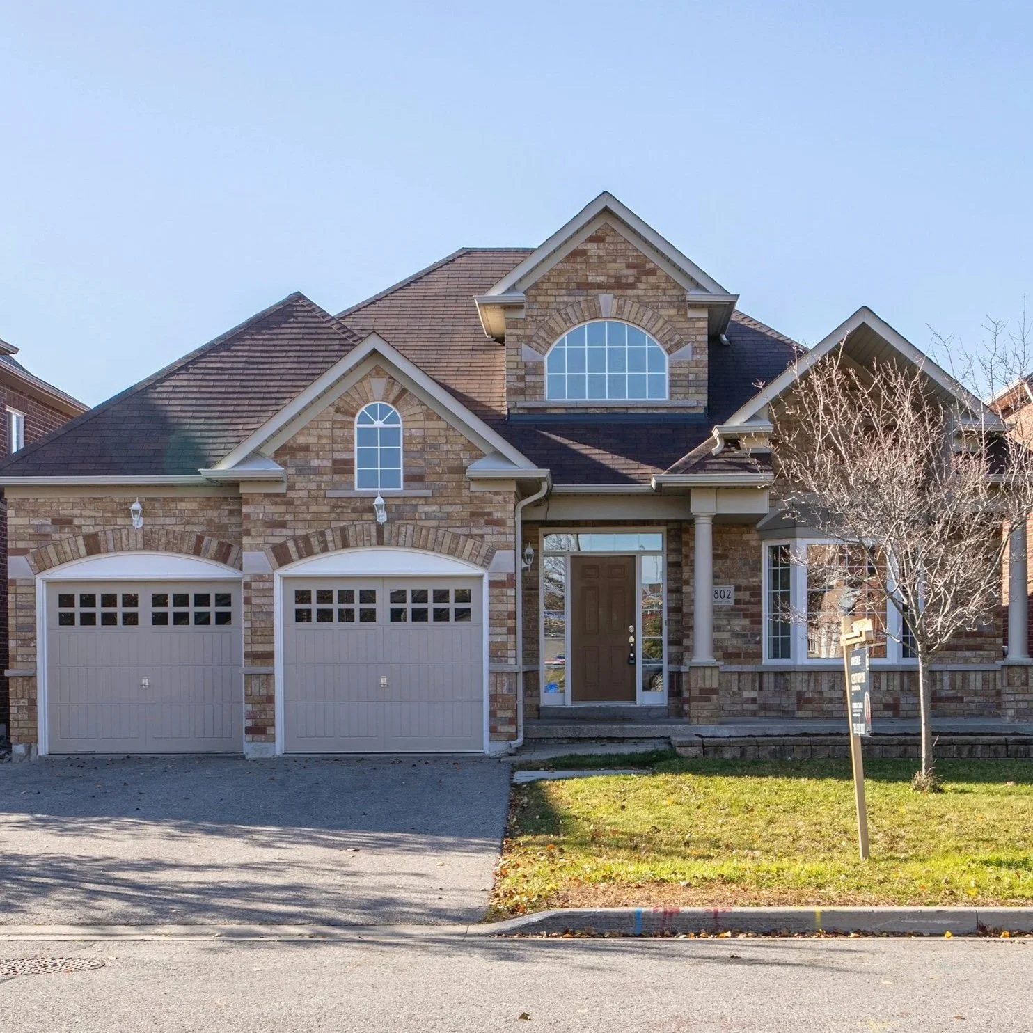 A two-story brick house with a front porch, two garage doors, and a large arched window above the entrance. There is a tree on the right side and a lawn in front.