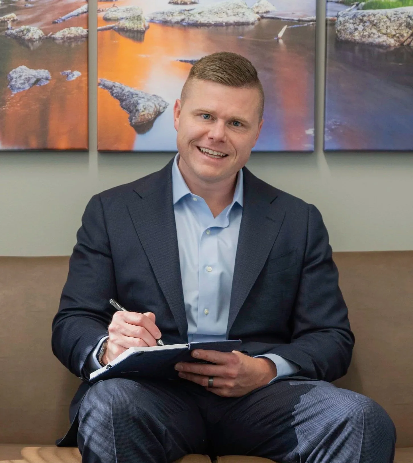Jonathan Thomas in a suit sitting on a bench, smiling, holding a pen and a notebook, with abstract landscape artwork on the wall behind him.