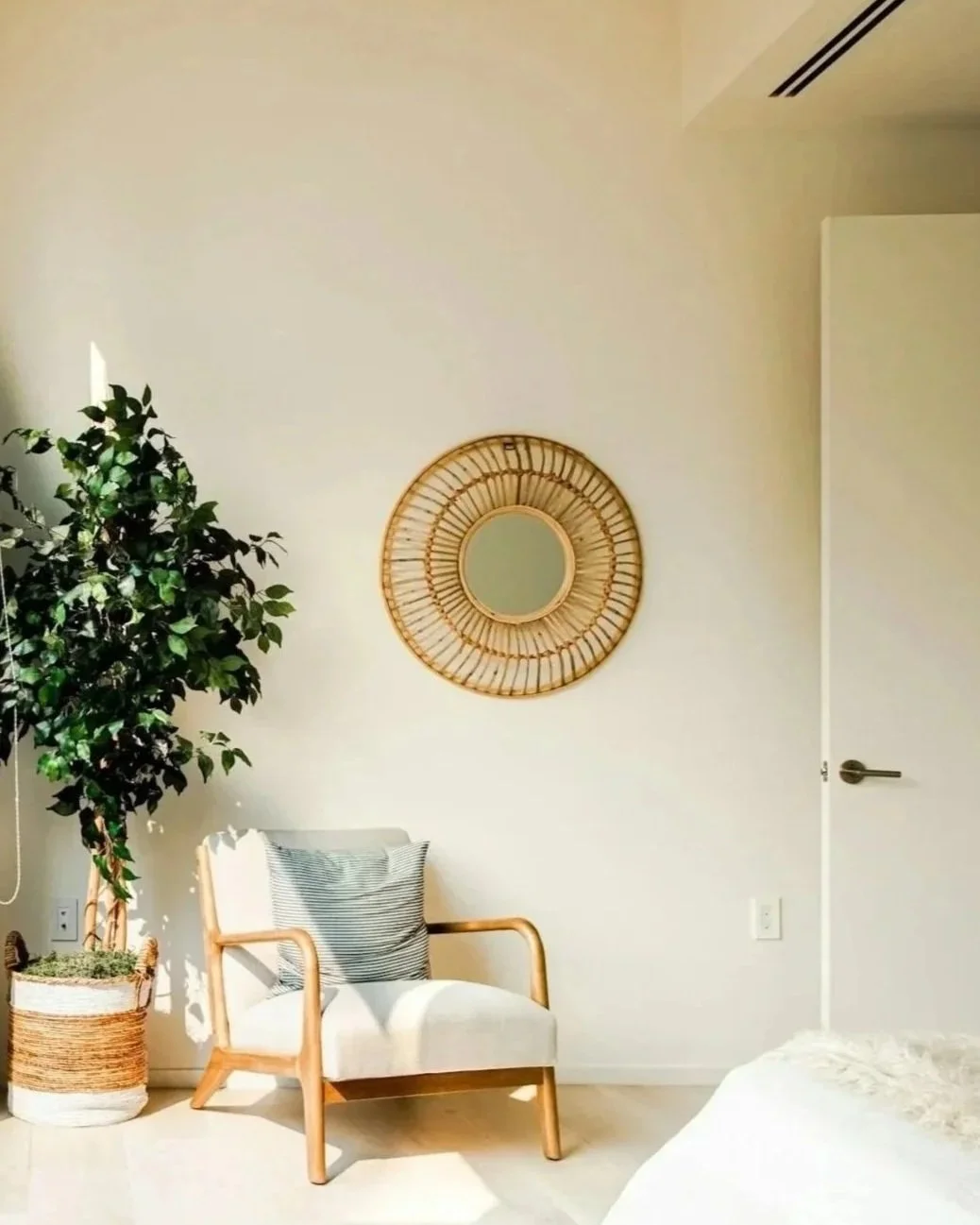 A minimalist interior room featuring a potted plant, a wicker mirror on the wall, a chair with a striped pillow, and a partially visible bed.