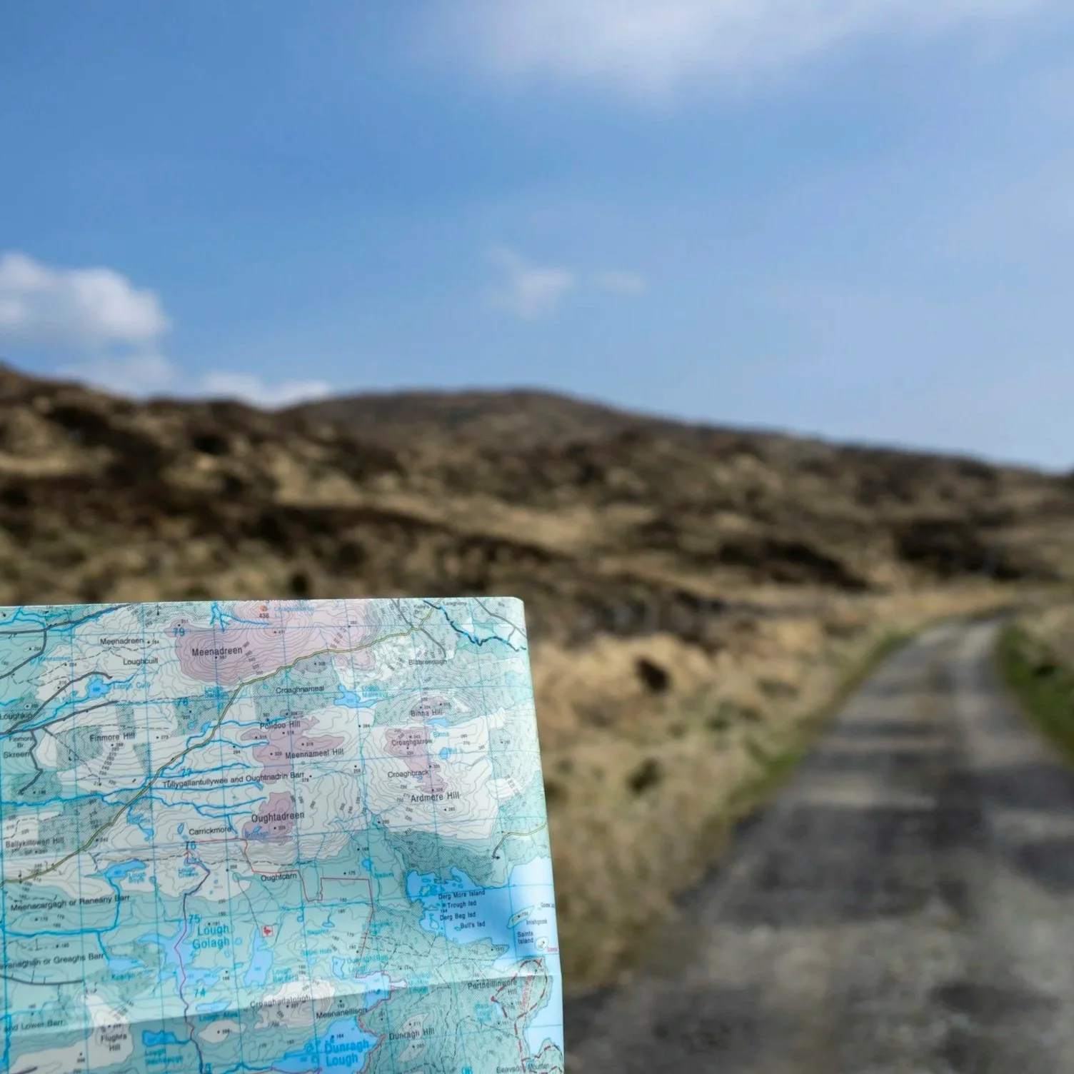 A map held in front of a winding trail in a hilly, rural landscape with brownish grass and a partly cloudy sky.
