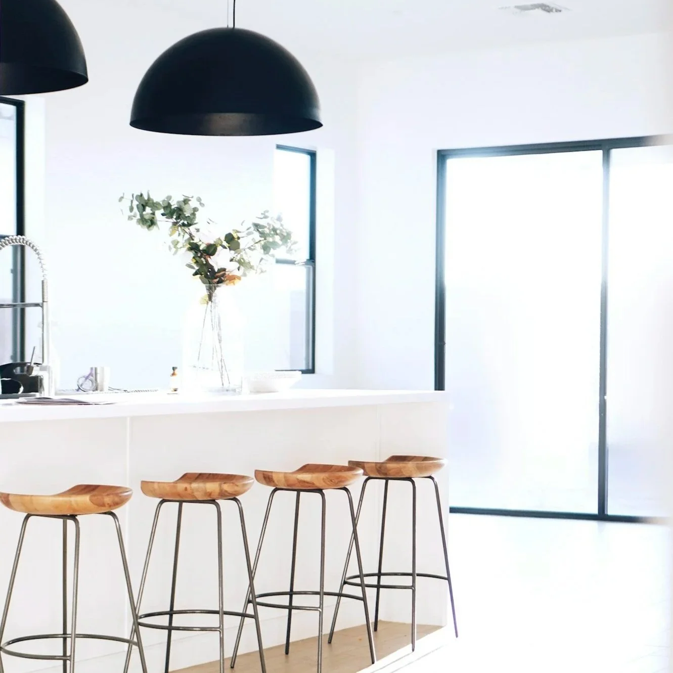 Bright, modern kitchen with black pendant lights, a white countertop island with four wooden bar stools, a vase with green leafy branches, and large windows with black frames.