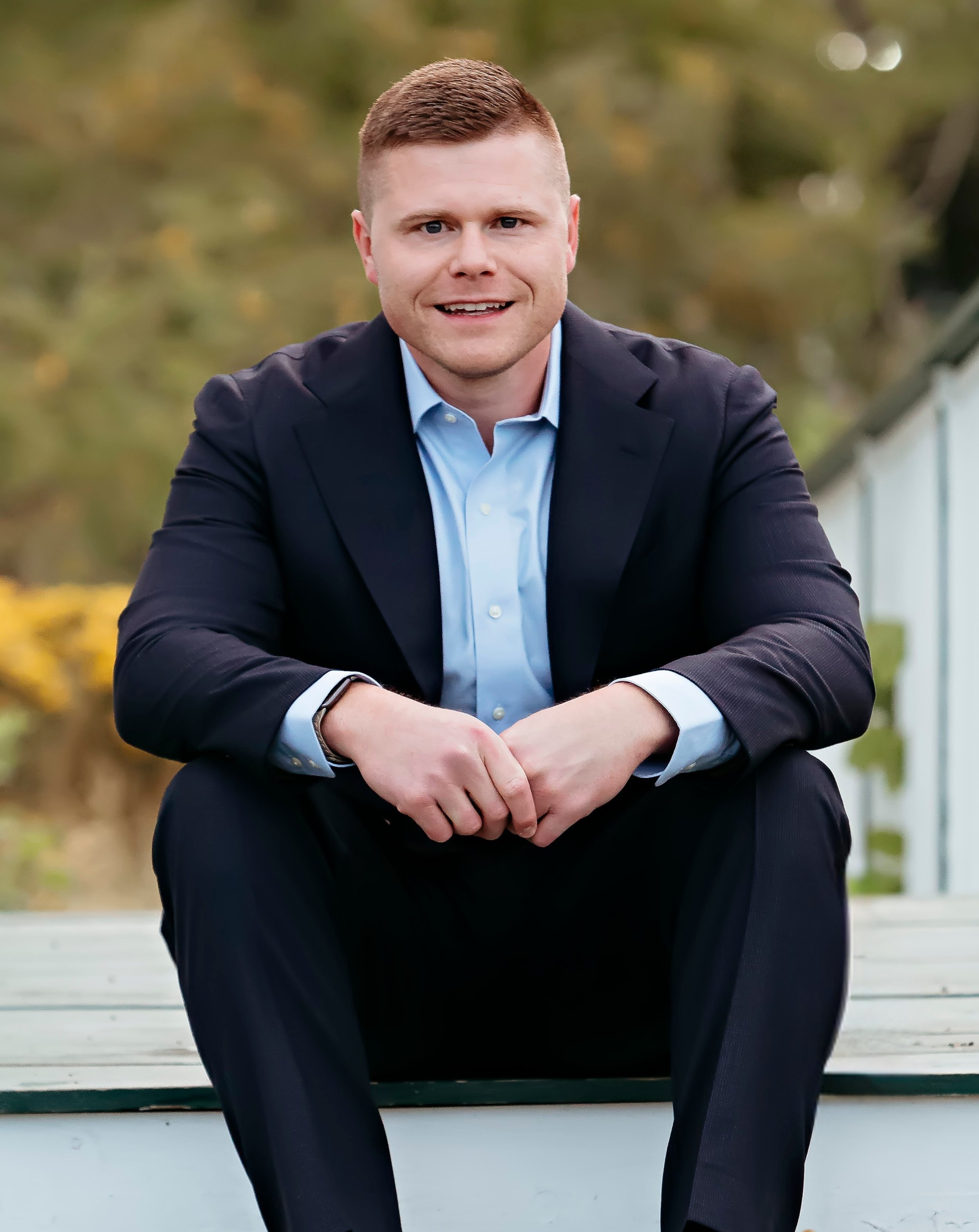 Jonathan Thomas in a navy suit and light blue shirt sitting outdoors on steps, with autumn trees in the background.