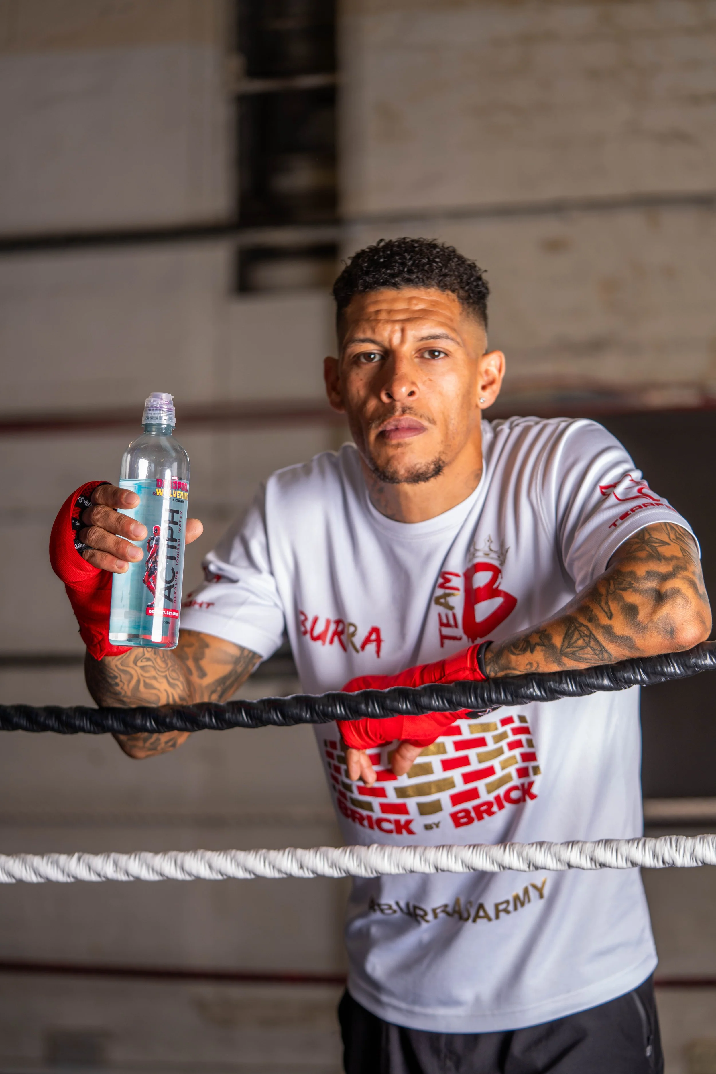 A man with tattoos and wearing boxing gloves and a white team shirt holding a water bottle inside a boxing ring.