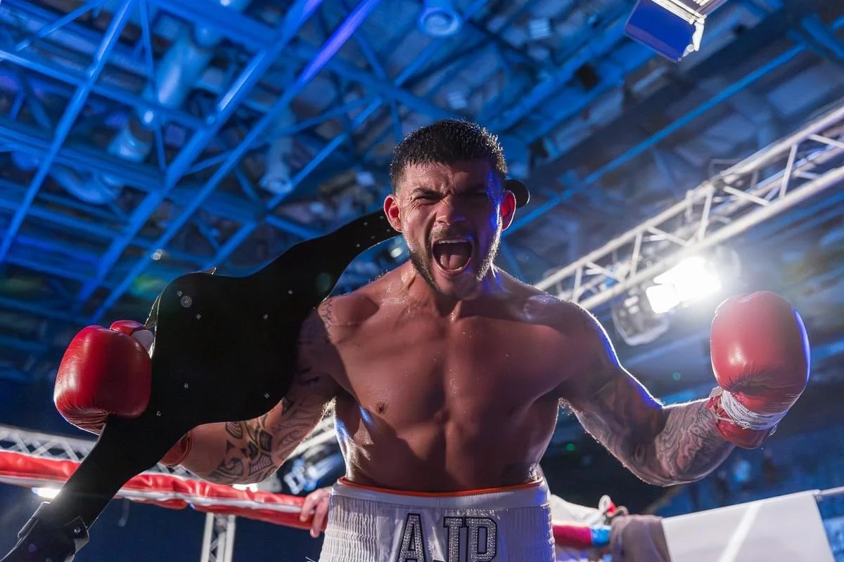 Boxer celebrating in the boxing ring after a match, holding his championship belt over his shoulder, with a triumphant expression.