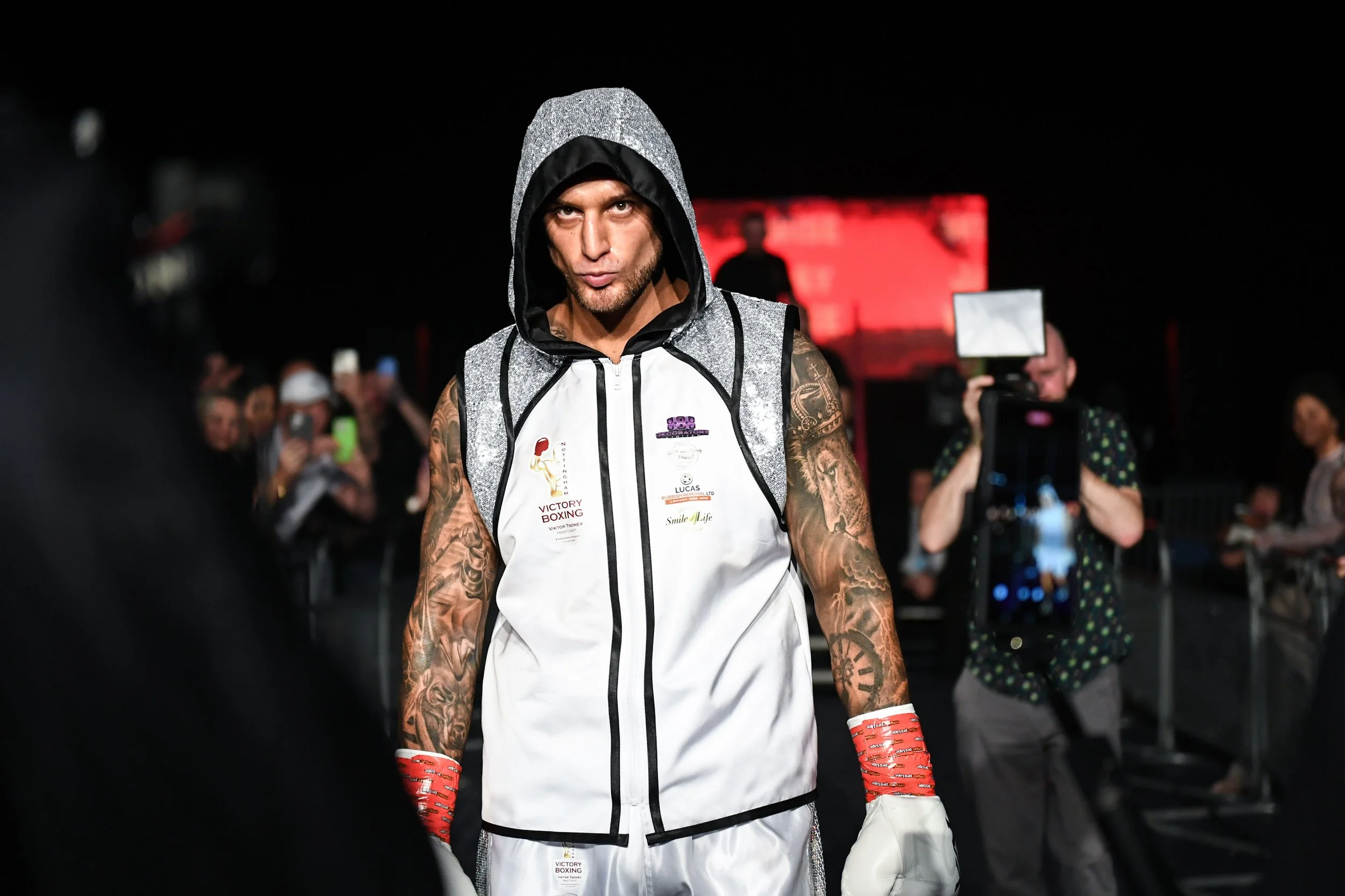 A male boxer with tattoos wearing a sleeveless hoodie and gloves, walking in a boxing arena with spectators and photographers in the background.
