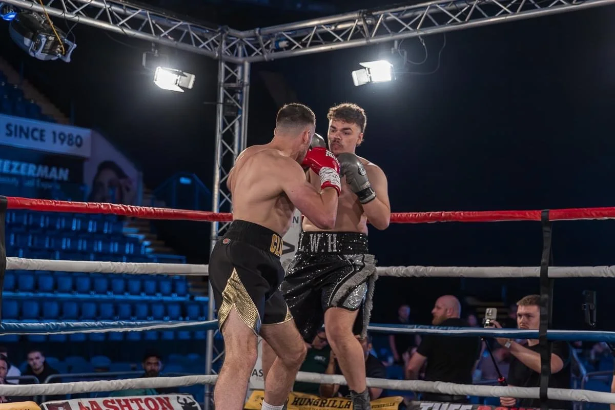 Two shirtless male boxers fighting in a boxing ring with fighters and spectators in the background under bright lights.