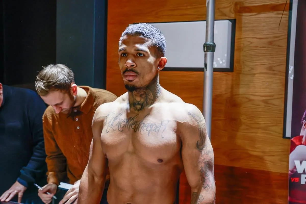 A shirtless man with tattoos standing in a room, possibly at a weigh-in or event, with two people behind him, one taking notes.