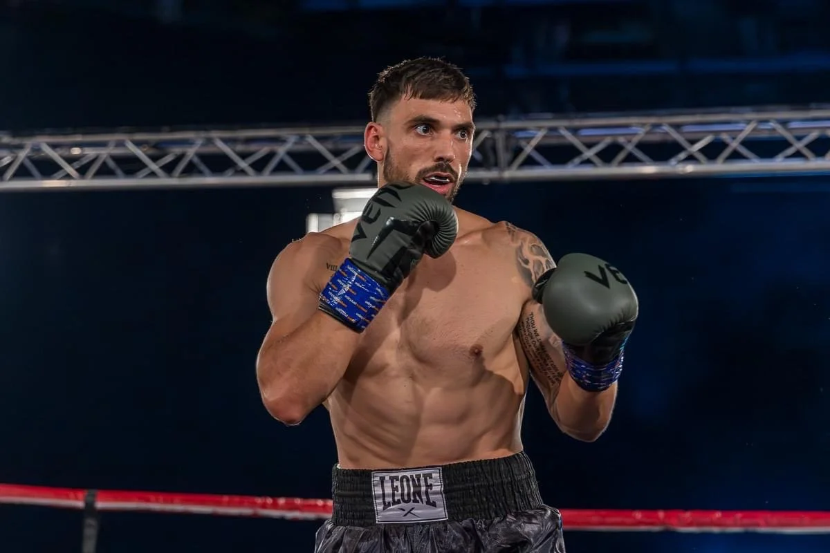 A male boxer in a fighting stance wearing black gloves and shorts, standing inside a boxing ring with a dark background and overhead lighting.
