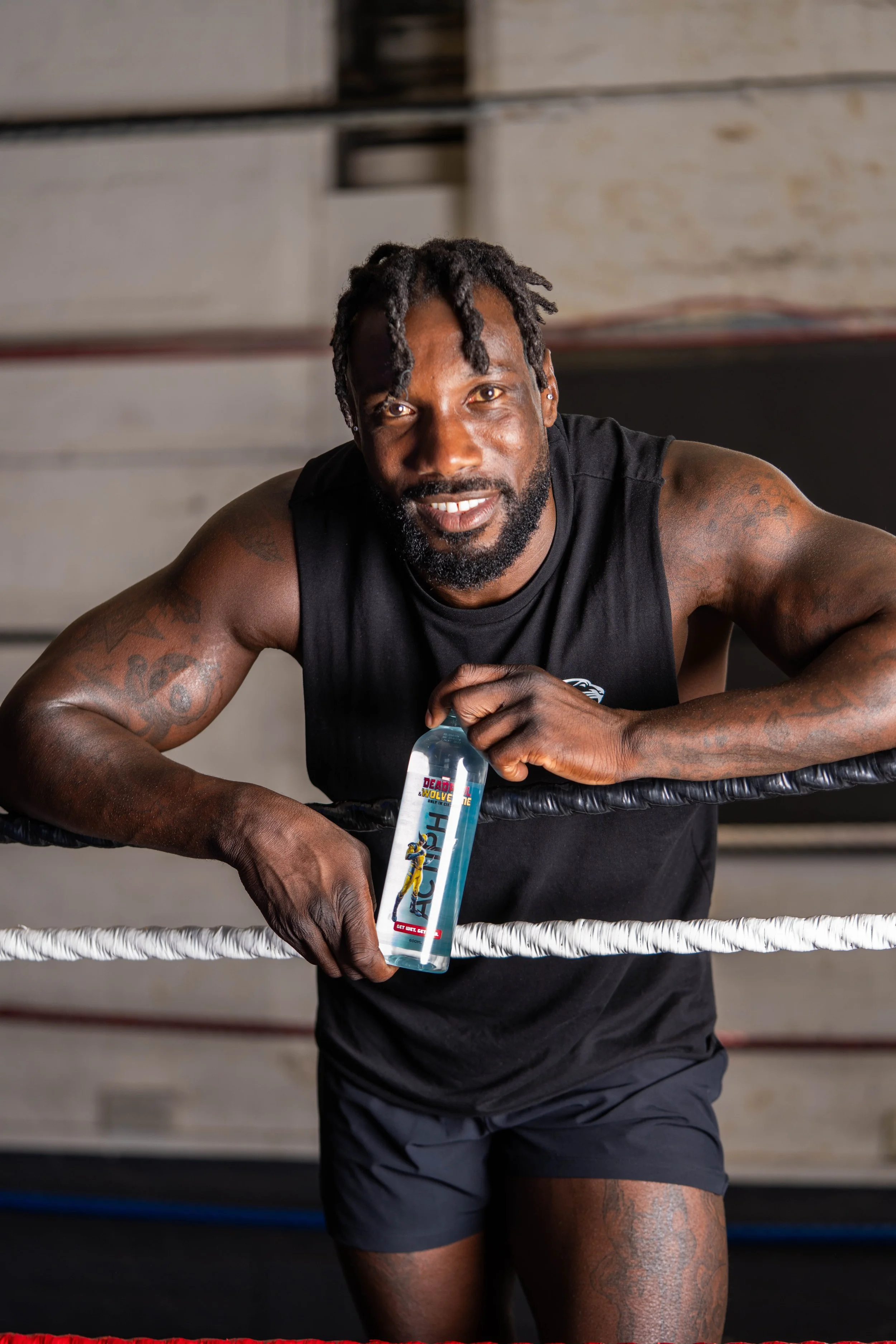 A man with tattoos on his arms in a boxing ring, smiling, holding a bottle of hand sanitizer labeled 'Death Battle' featuring a graphic of a character.