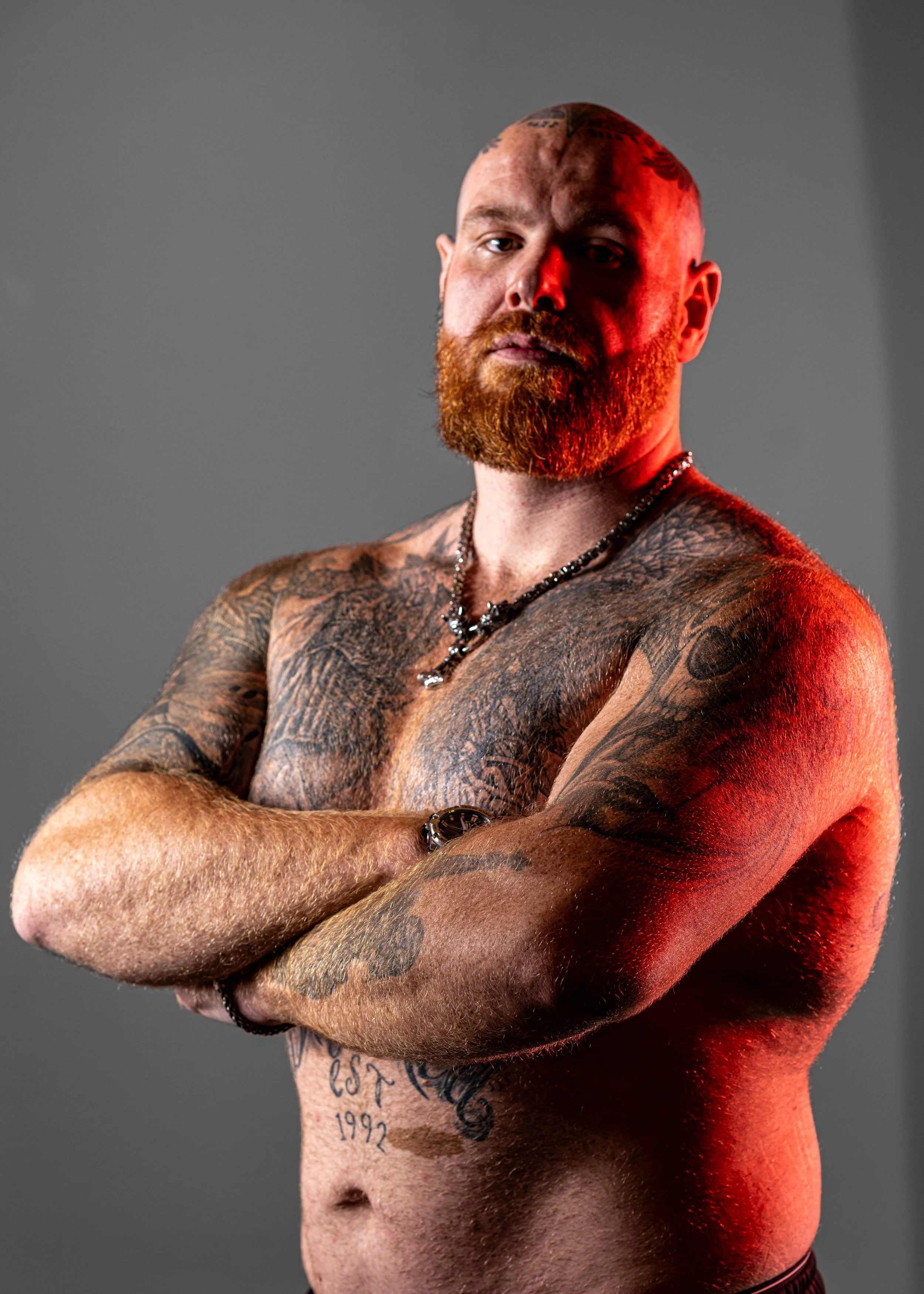 A shirtless man with tattoos on his arms and chest, crossing his arms, with a beard and a shaved head, standing against a gray background with red lighting on one side of his face and body.