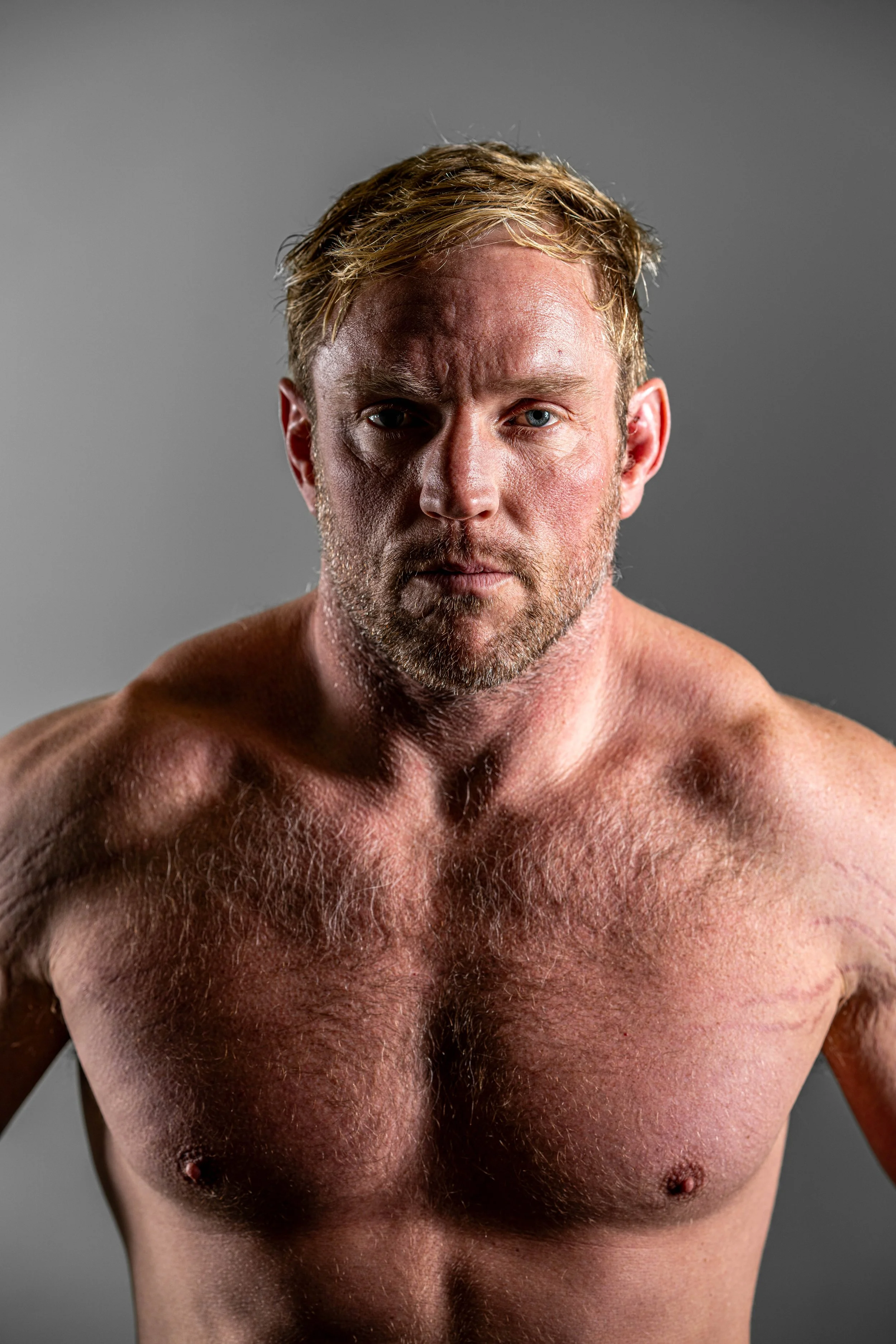 A shirtless man with short blond hair and a beard looking directly at the camera against a plain gray background.