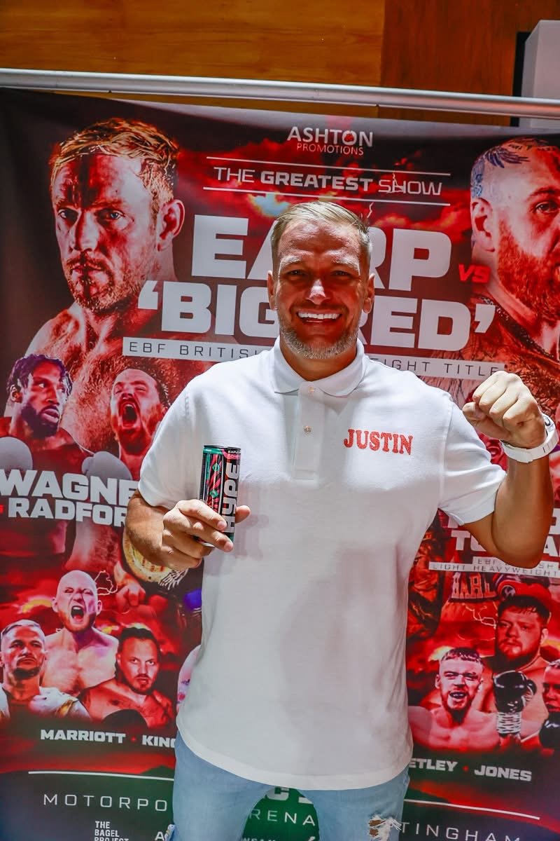 A man wearing a white polo shirt with the name 'Justin' stands in front of a promotional poster for a boxing event called 'The Greatest Show'. He is smiling, holding a can of energy drink in his right hand, and raising his left fist in a victory gesture. The poster features images of various boxers and event details.