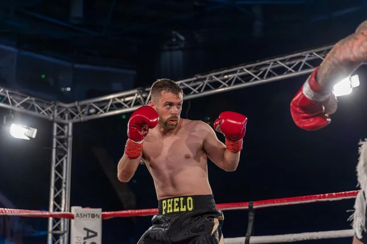 A shirtless male boxer wearing red gloves and black shorts with his name 'PHELO' on the waistband boxs in a boxing ring.