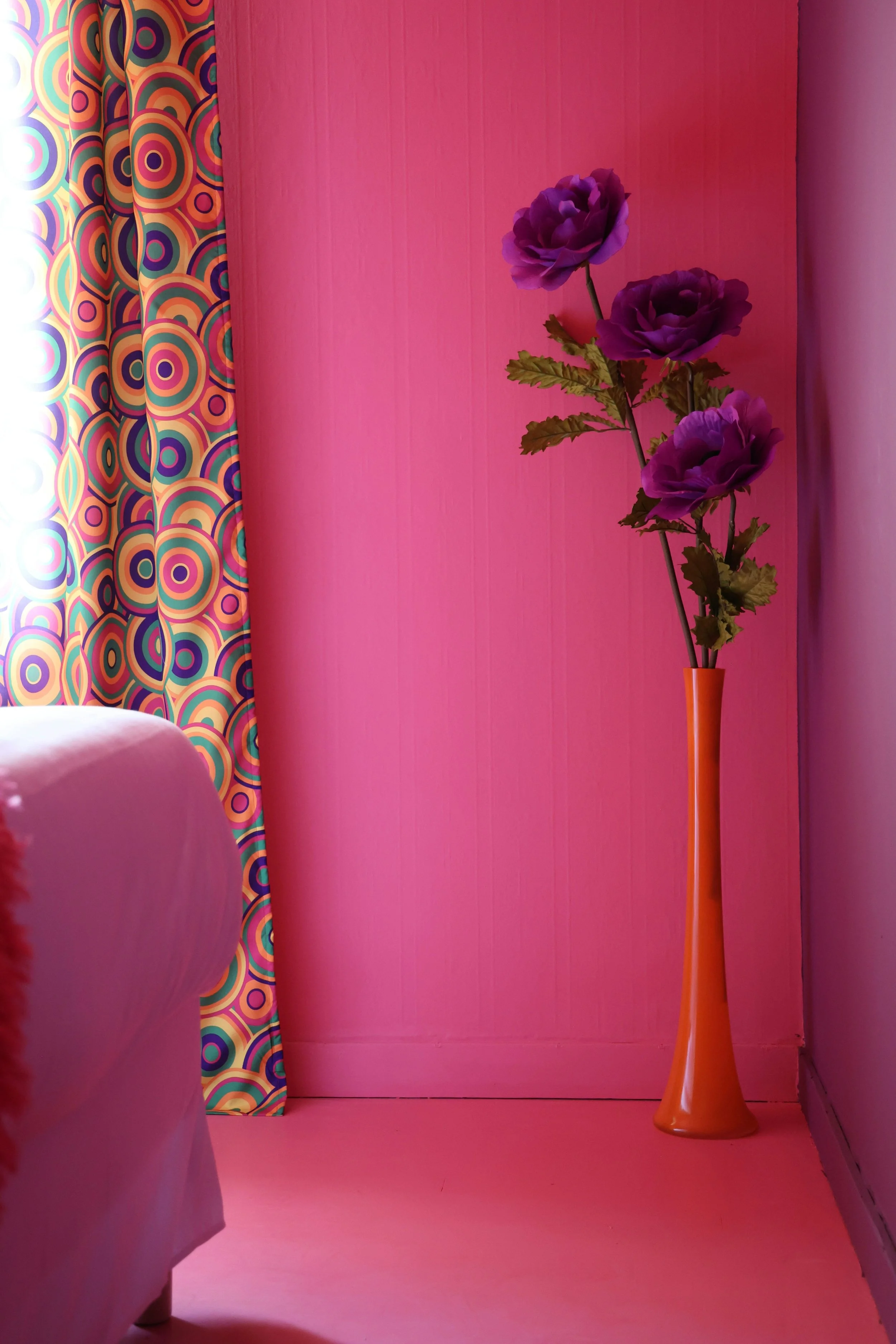 Pink room corner with colorful circular patterned curtains, pink wall, and a tall orange vase with purple artificial flowers.