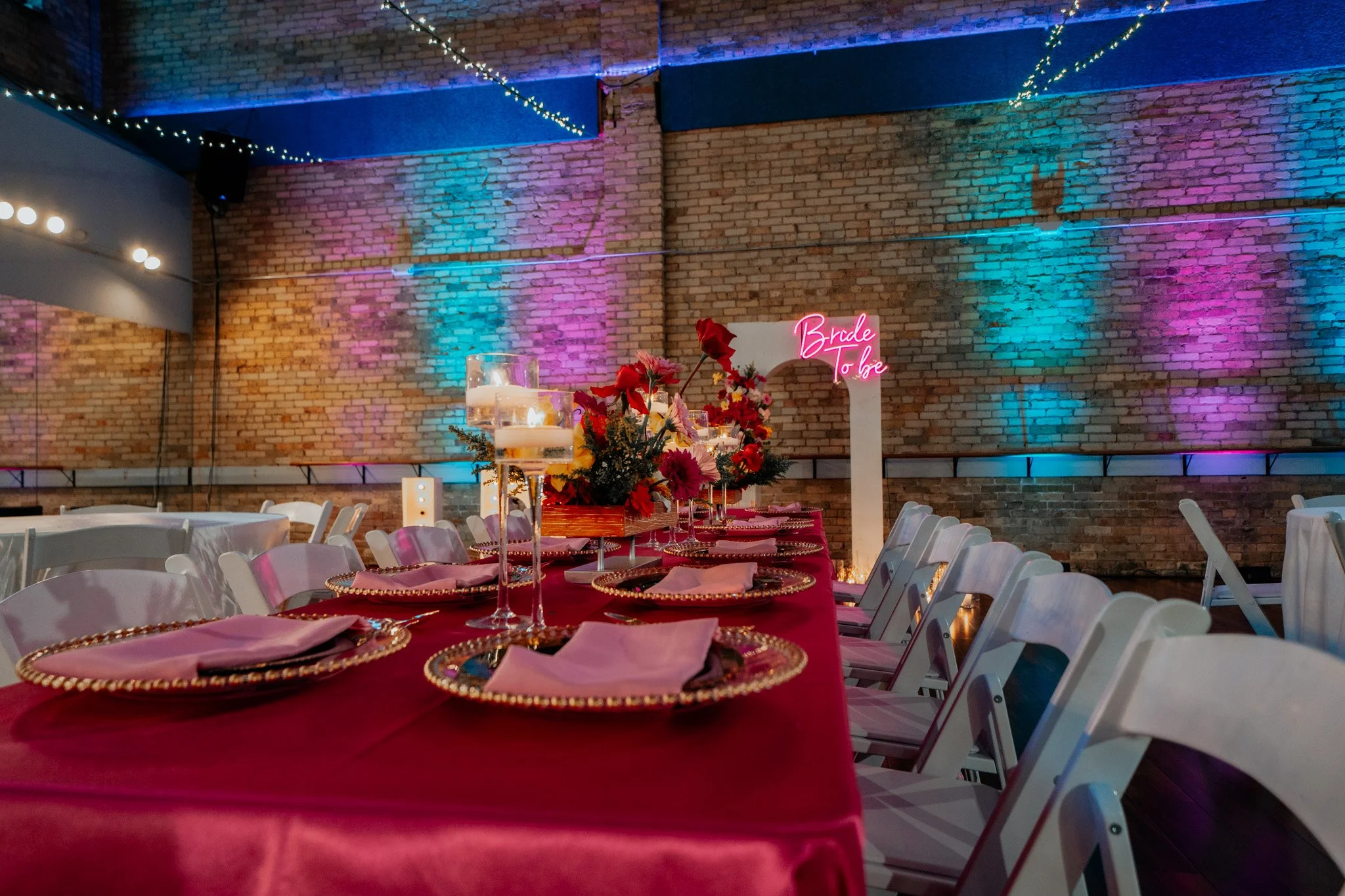 Decorated event table with pink tablecloth, floral centerpiece, hanging candles, and a neon sign that says 'Bride to be' against a brick wall with colorful uplighting.