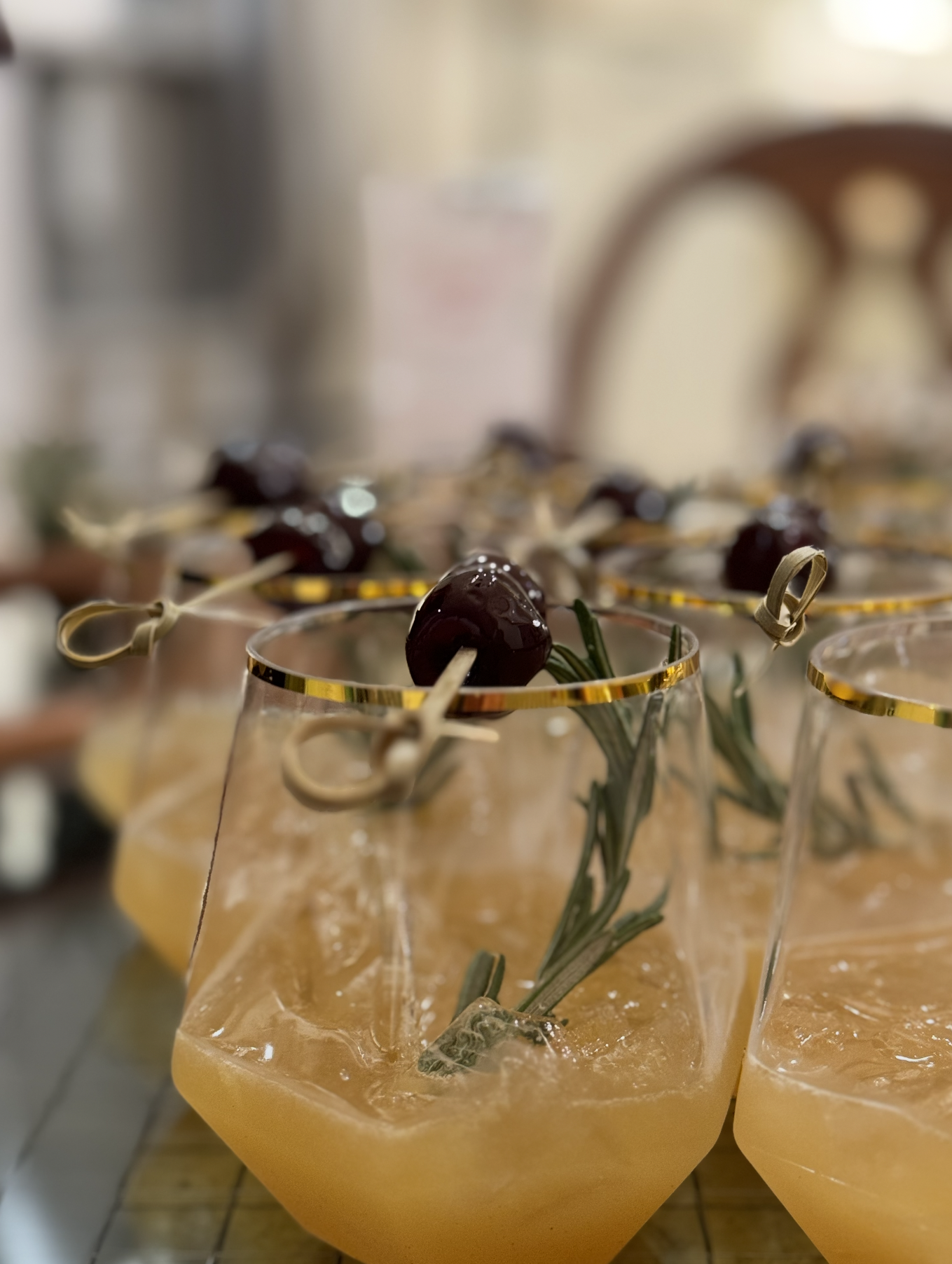Close-up of multiple glasses filled with a yellow-orange beverage, garnished with a sprig of rosemary and topped with a cherry, with a blurred background.