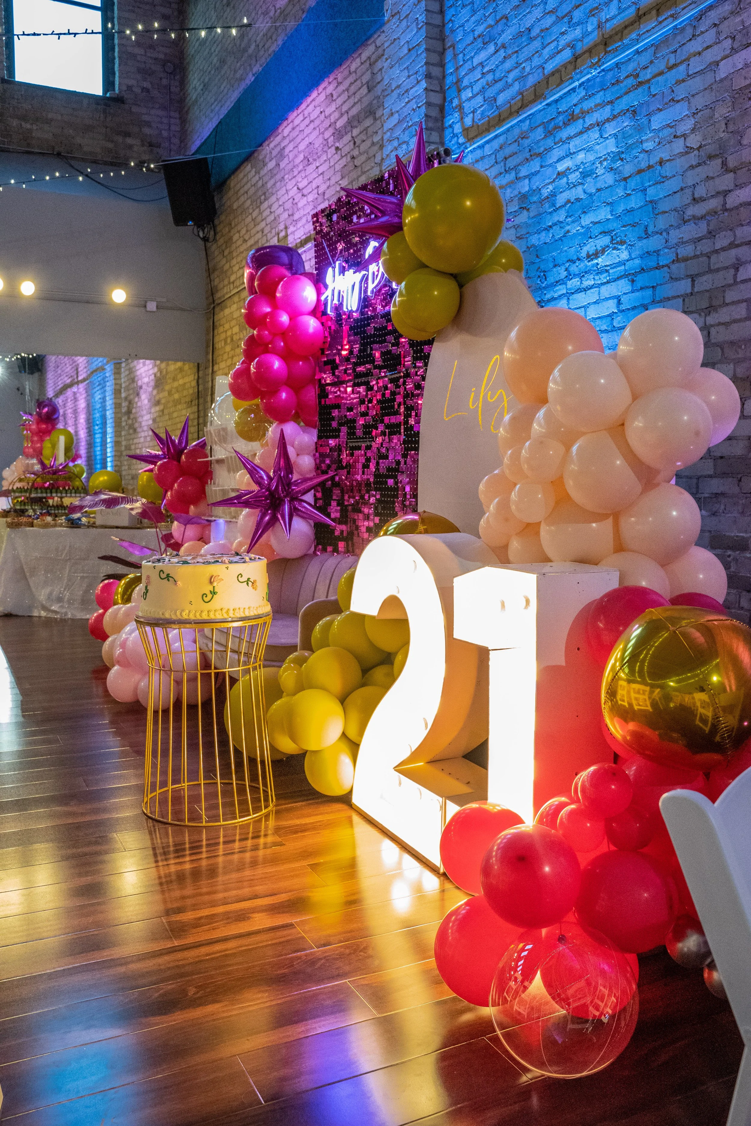 Decorations for a 21st birthday celebration featuring colorful balloons in pink, yellow, and gold, large illuminated "21" signs, and a backdrop decorated with pink, purple, and gold stars, set in an industrial-style venue with exposed brick walls and hardwood floors.