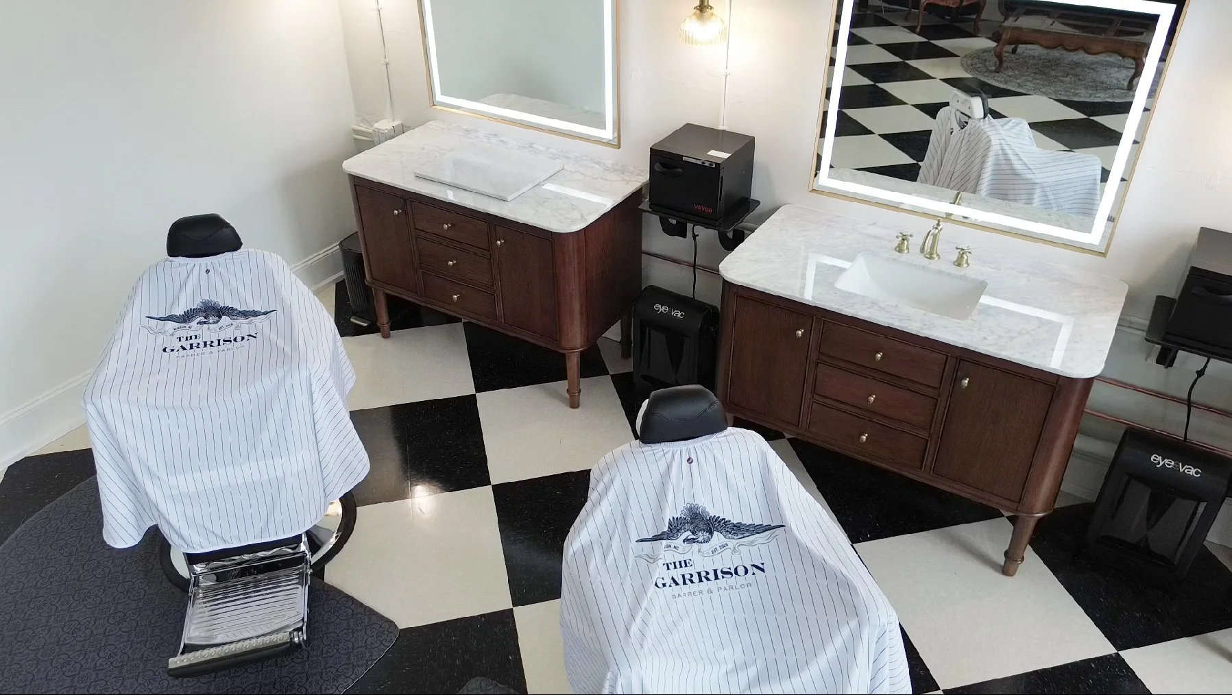 Barber shop with two chairs covered with striped capes, mirrors, and two wooden counters with marble tops, black and white checkered floor.