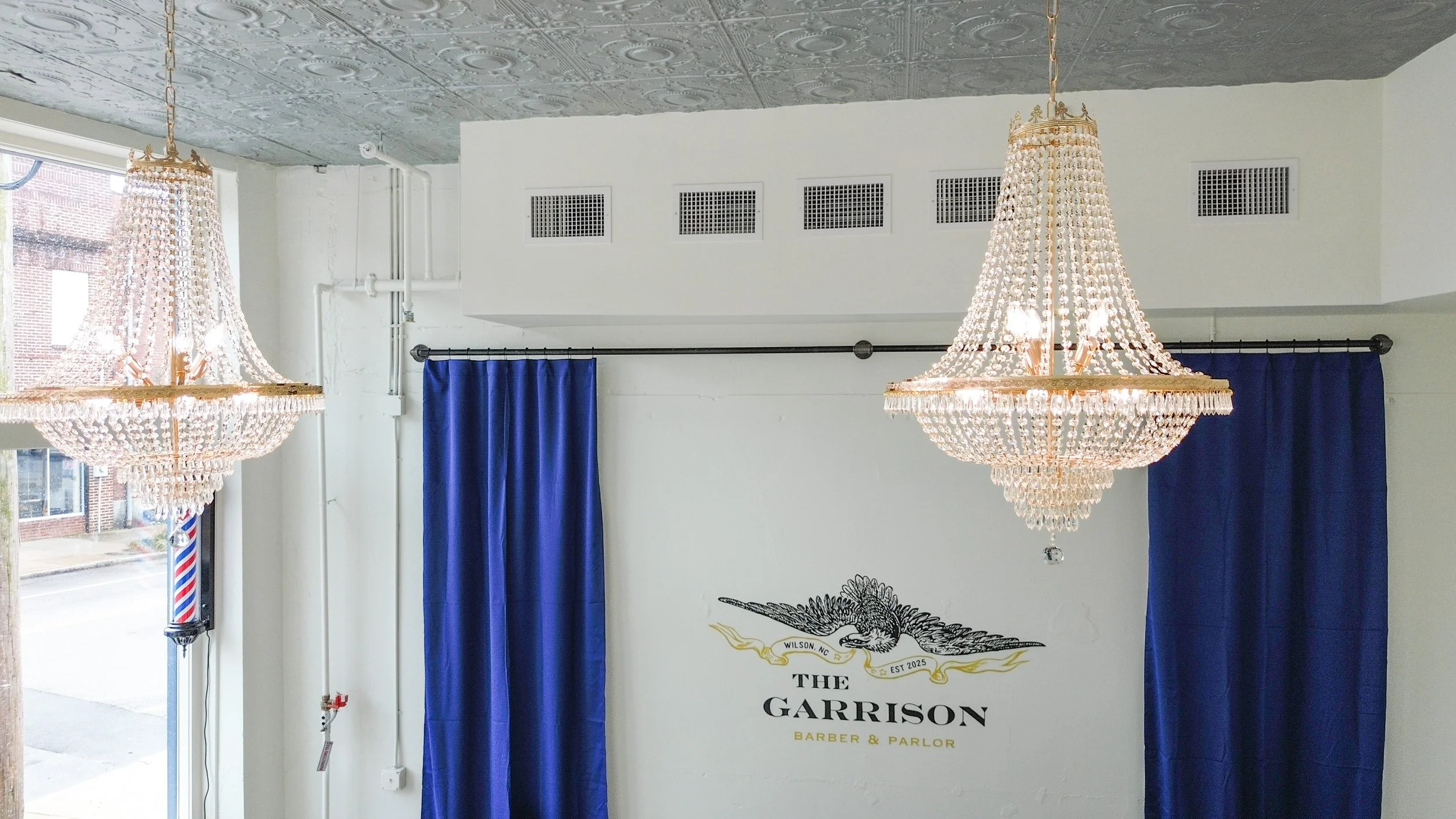 Interior of a barber shop named The Garrison, featuring two chandeliers hanging from the ceiling, blue curtains on the windows, and a wall with a logo of an eagle and the shop's name.