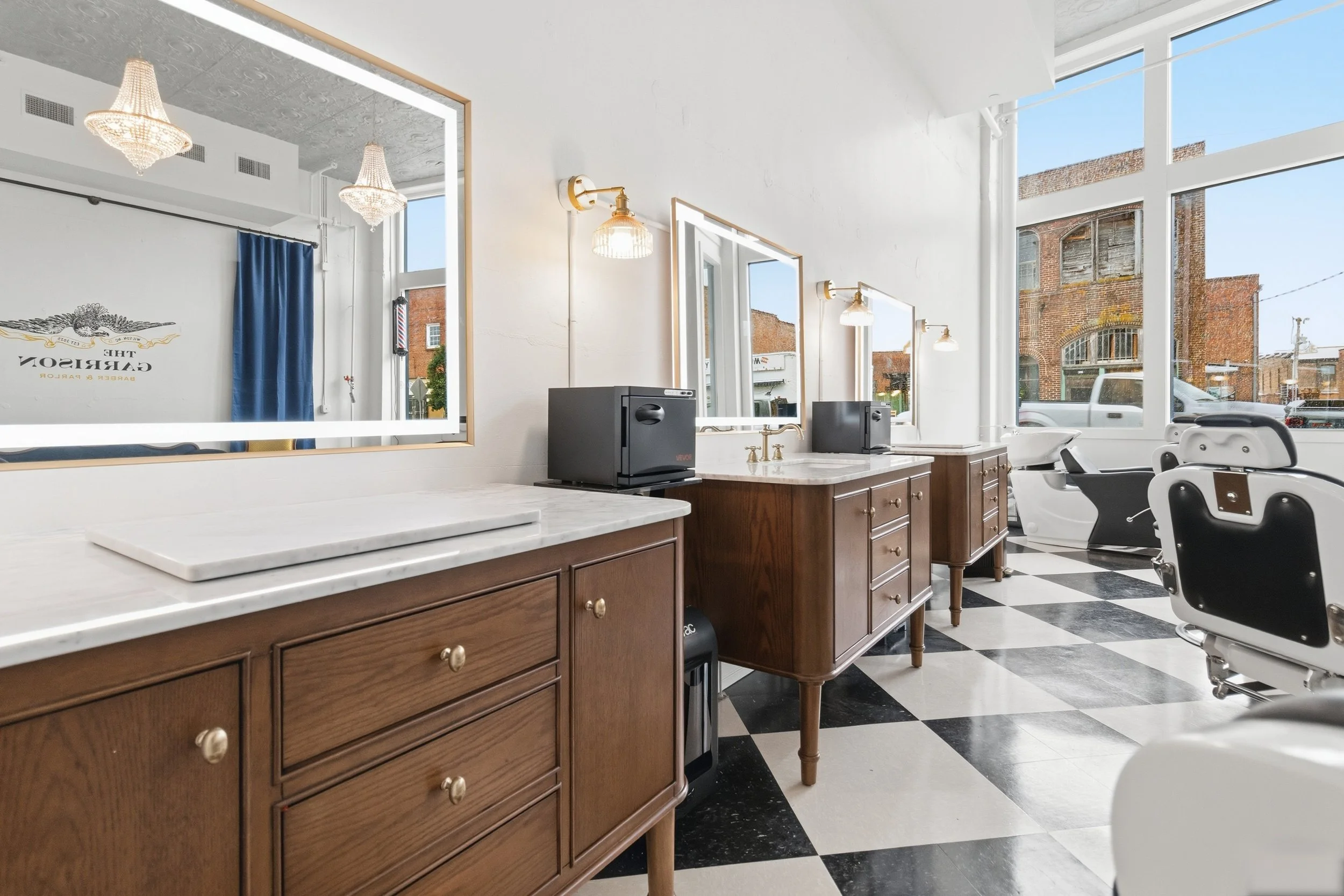 Interior of a vintage barber shop with wooden cabinets, mirrors, barber chairs, large windows, and checkered flooring.