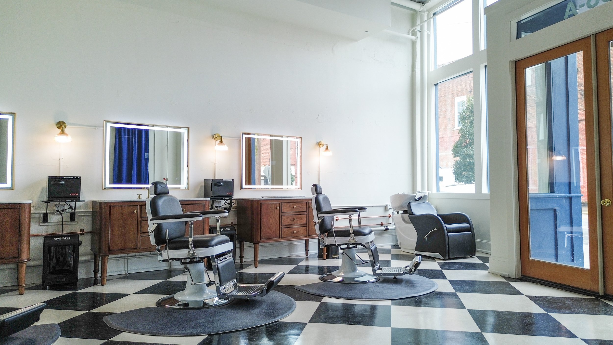 Modern barber shop with black and white checkered floor, two barber chairs, mirrors, mirrors, and natural light through large windows.