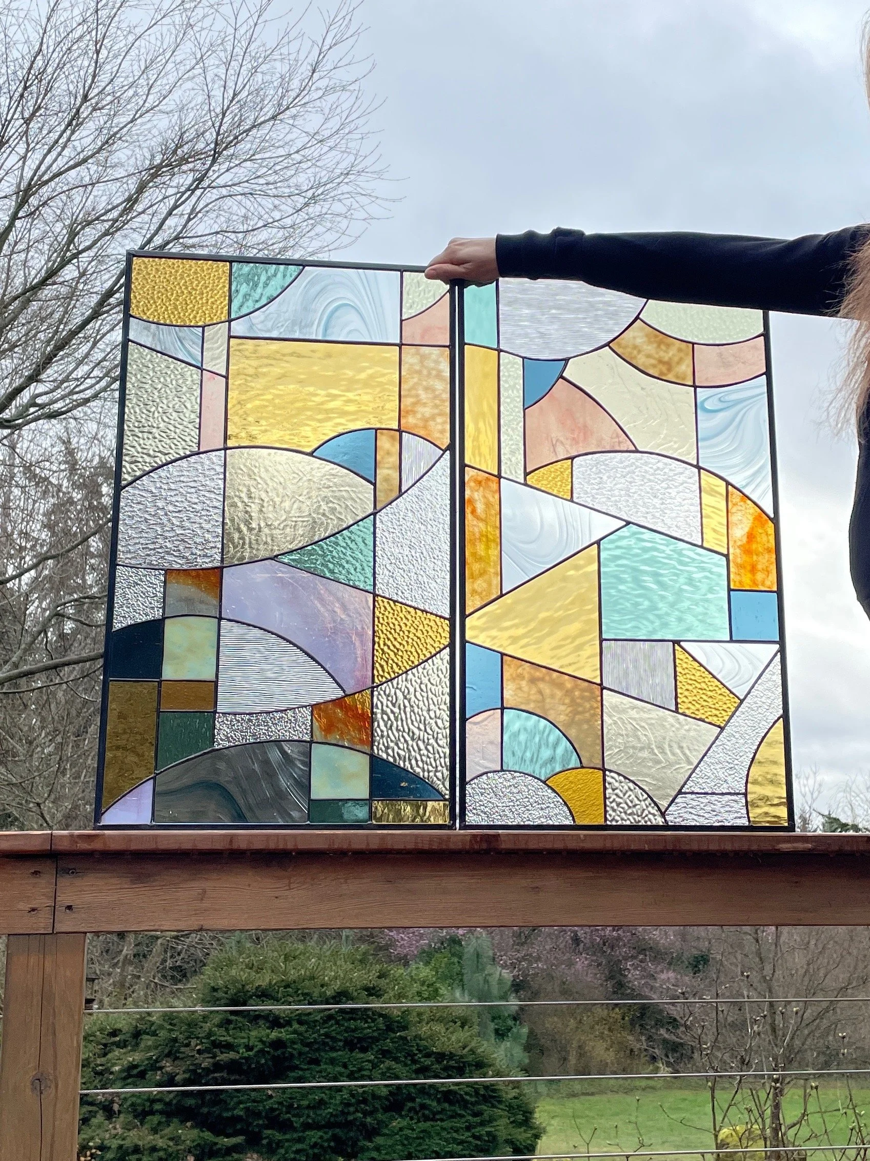 Colorful stained glass panel with abstract geometric patterns held up outside on a wooden railing, with trees and a cloudy sky in the background.