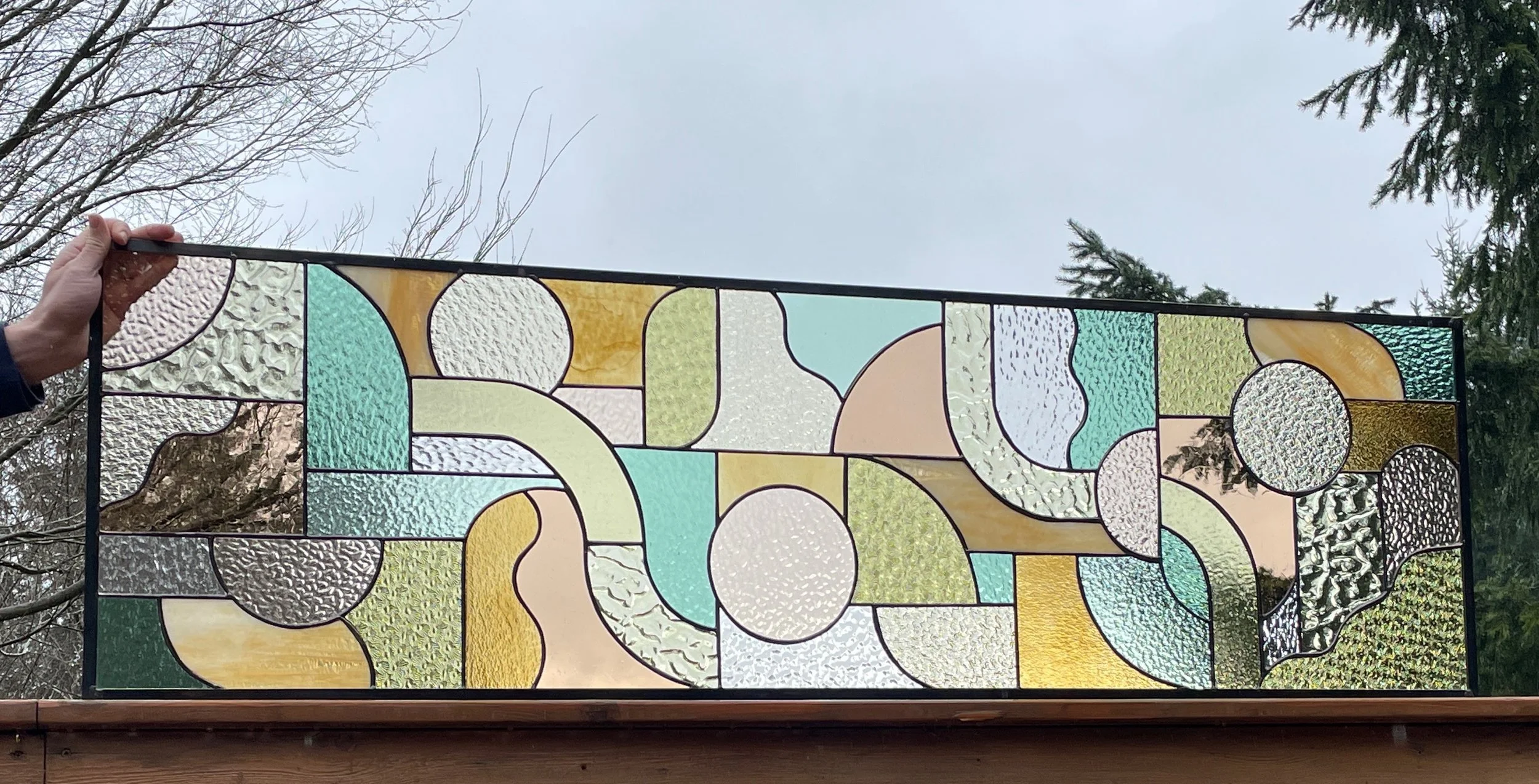 A rectangular stained glass window with abstract geometric shapes in pastel colors, held by a person's left hand against an outdoor sky and trees.