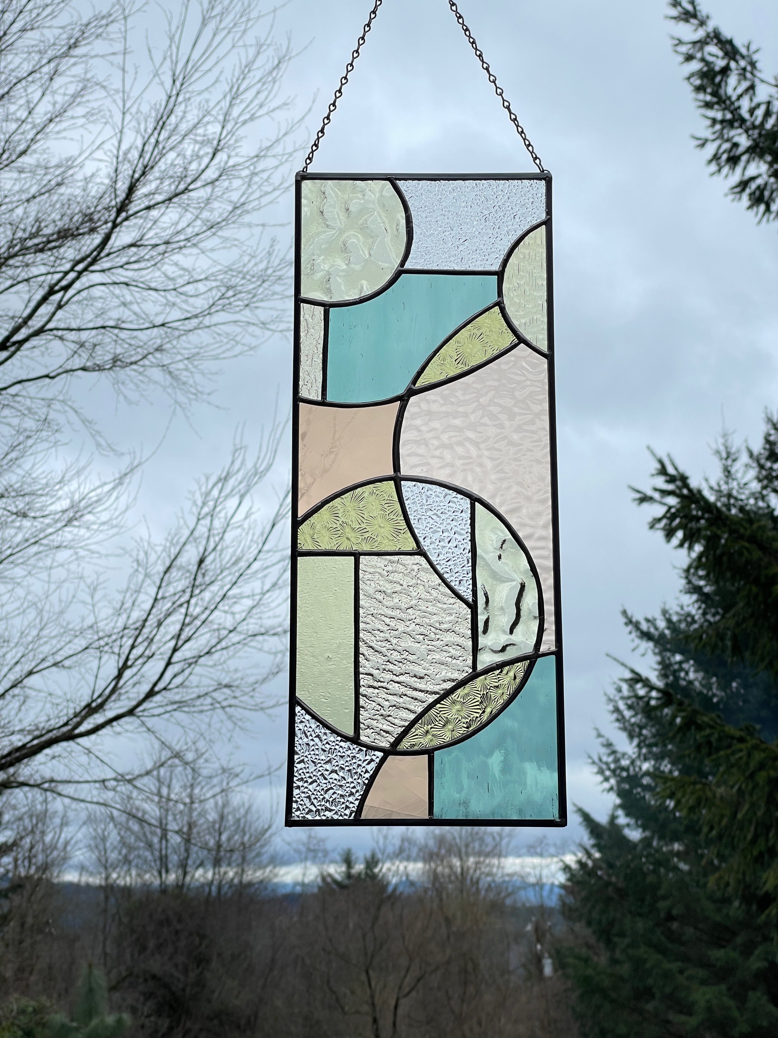 A hanging stained glass art piece with geometric patterns, featuring textured and colored glass in shades of blue, green, beige, and clear, suspended outdoors against a cloudy sky and trees.