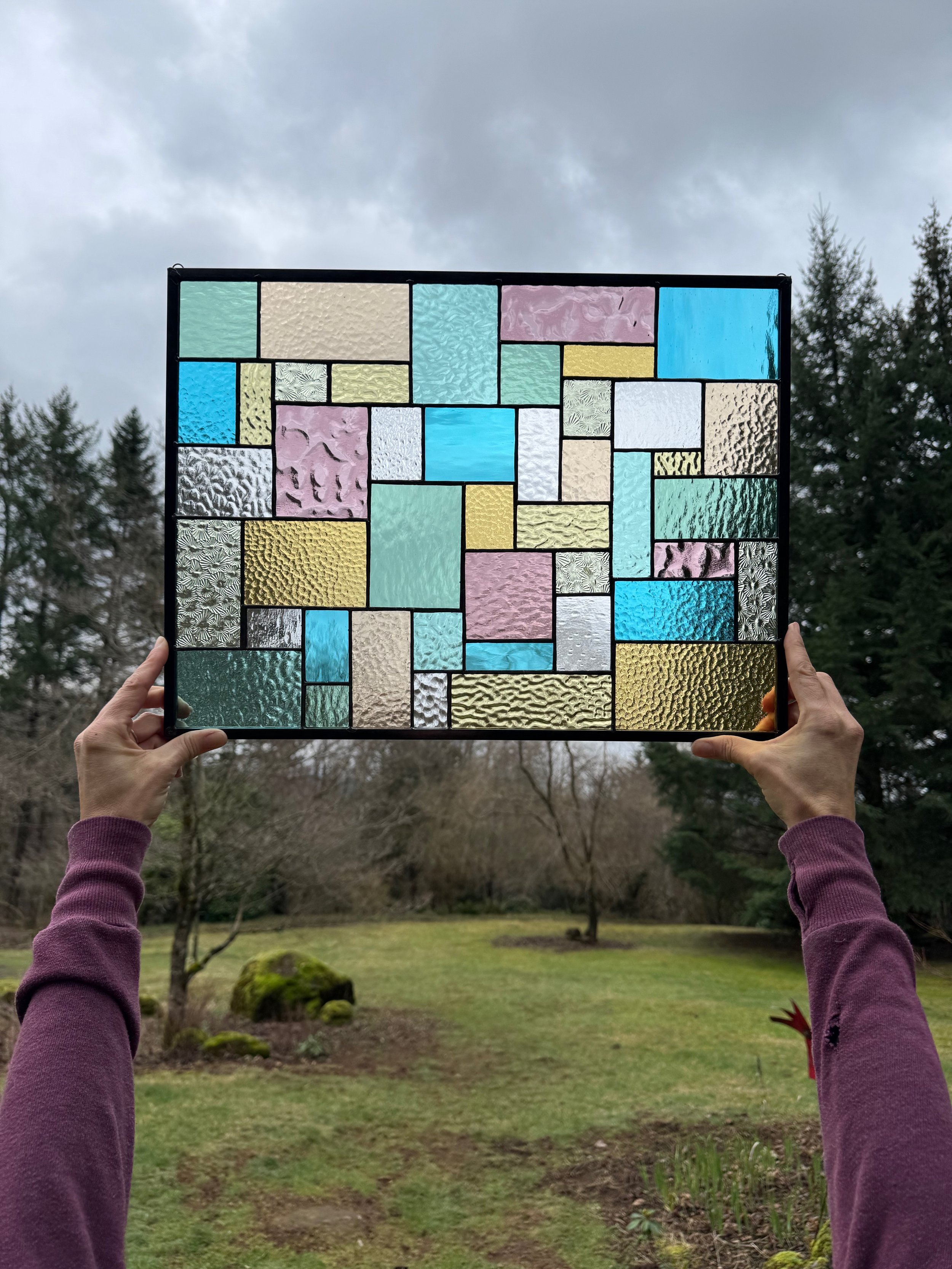 Person holding a stained glass window with multicolored rectangular glass pieces outdoors on a cloudy day.