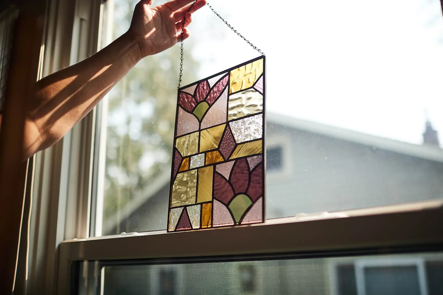 A hand hanging a stained glass art piece with floral and geometric patterns by a window, sunlight illuminating the colored glass.