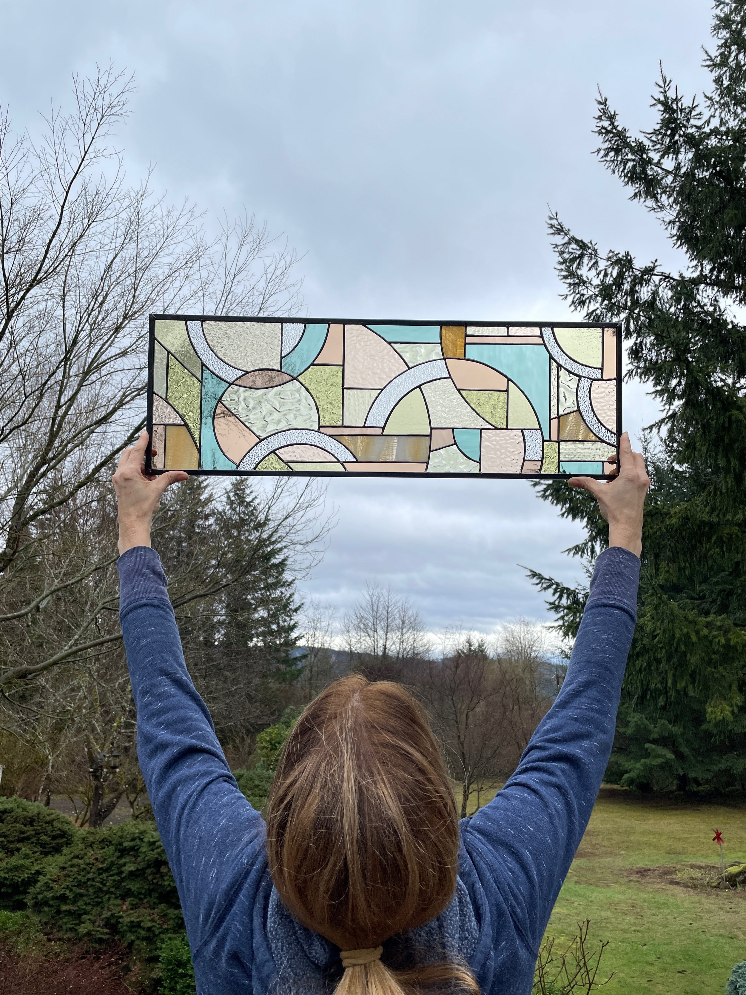 Person holding up a colorful stained glass artwork outdoors with trees and cloudy sky in the background.