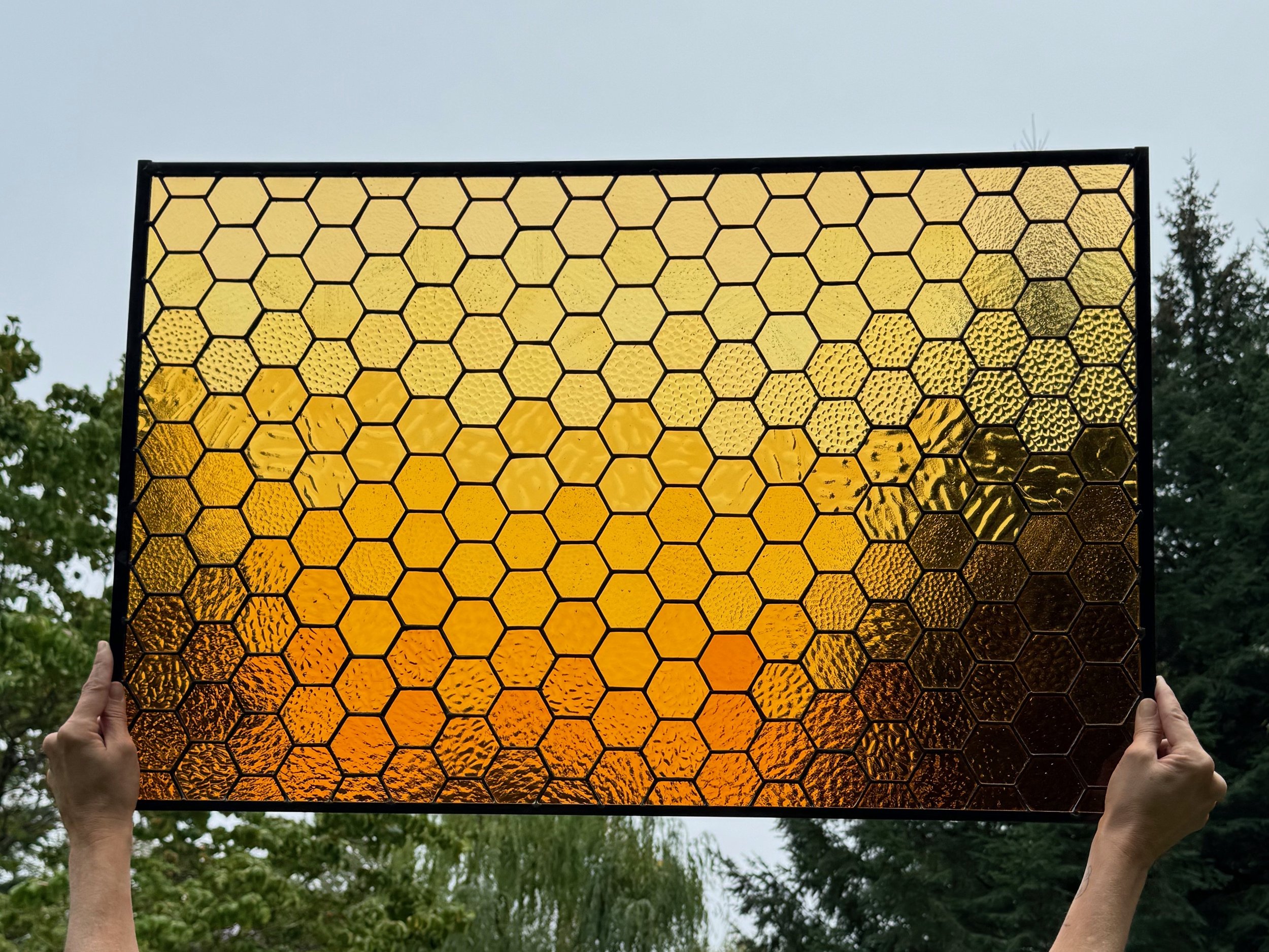 Person holding a large rectangular stained glass panel with a honeycomb pattern of various shades of yellow and orange, outdoors with trees in the background.