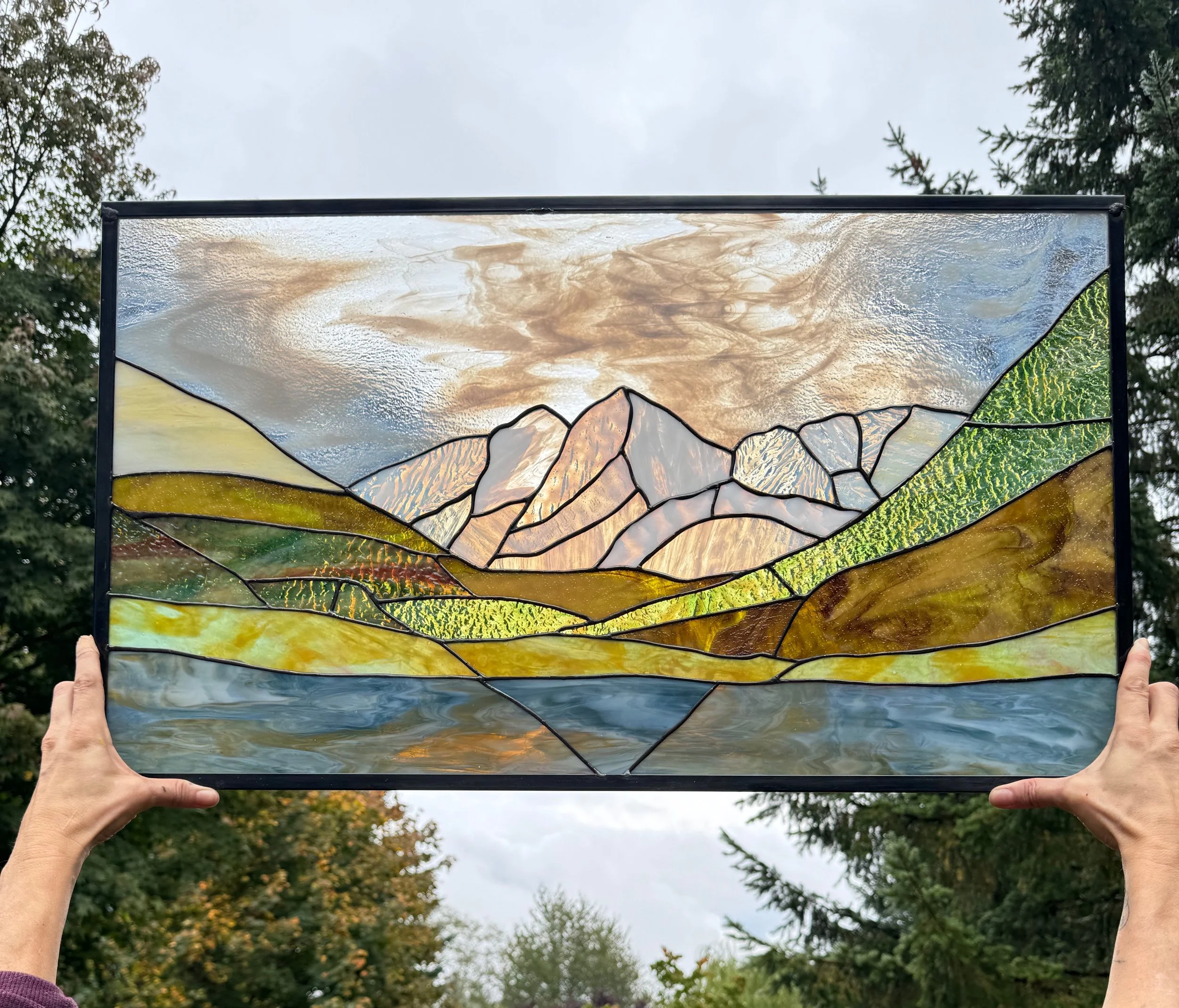 Person holding up a stained glass artwork depicting mountains, a river, and grassy hills under a cloudy sky.