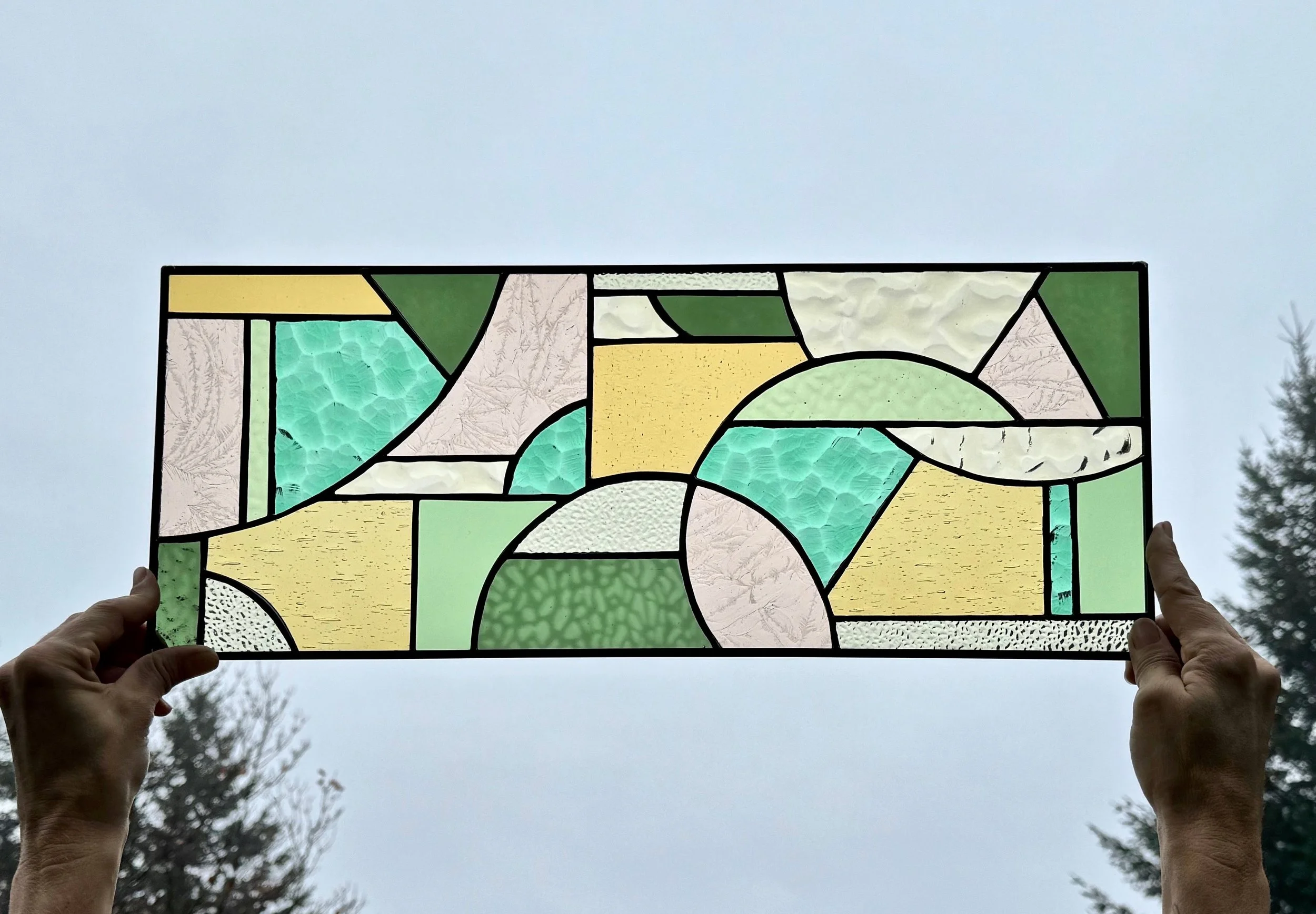 A person holds a stained glass window up to the sky, revealing an abstract design with various geometric shapes in shades of green, yellow, white, and clear textured glass.