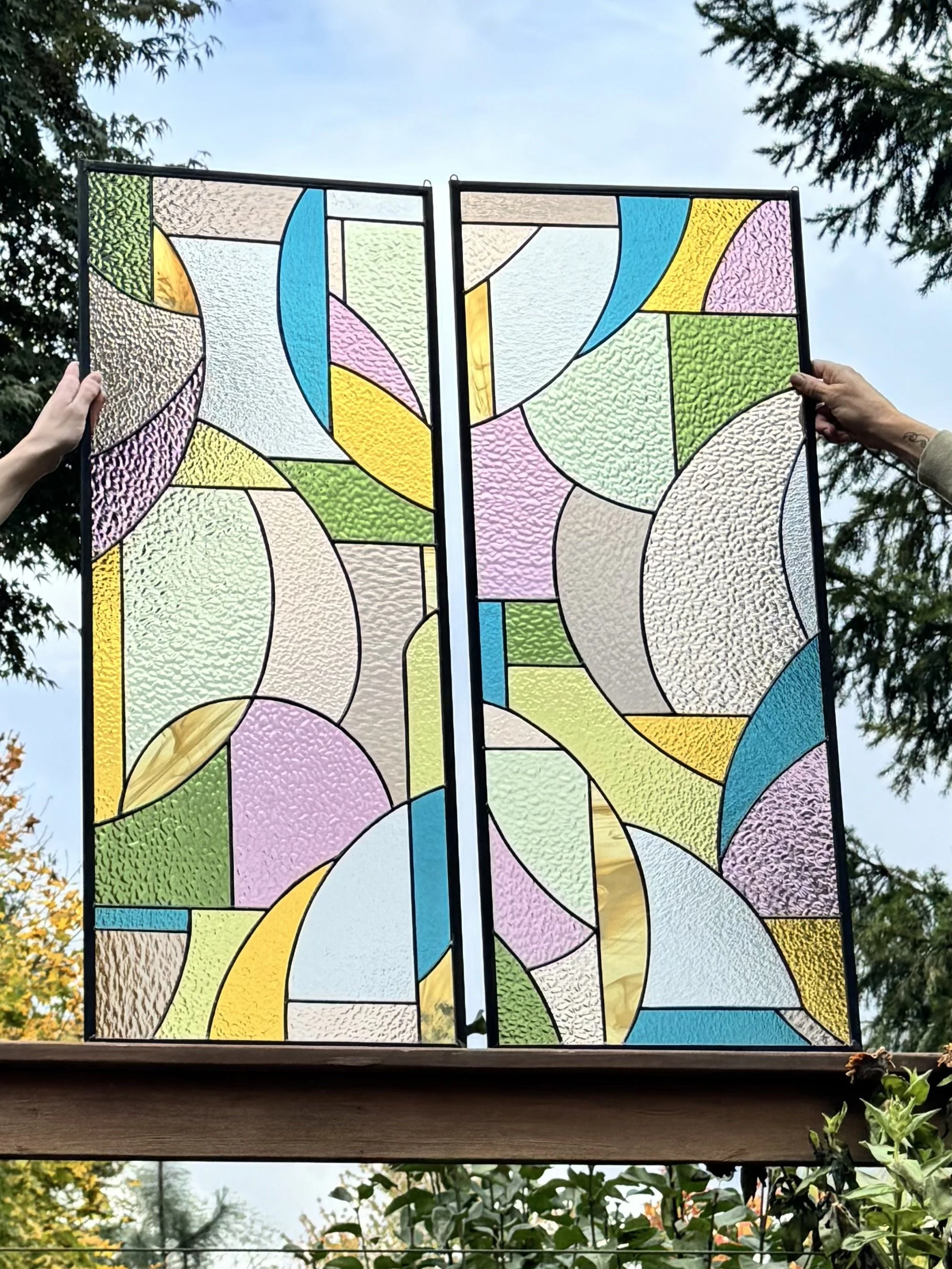 Colorful stained glass panel with abstract geometric shapes being held up outdoors against a sky and trees.