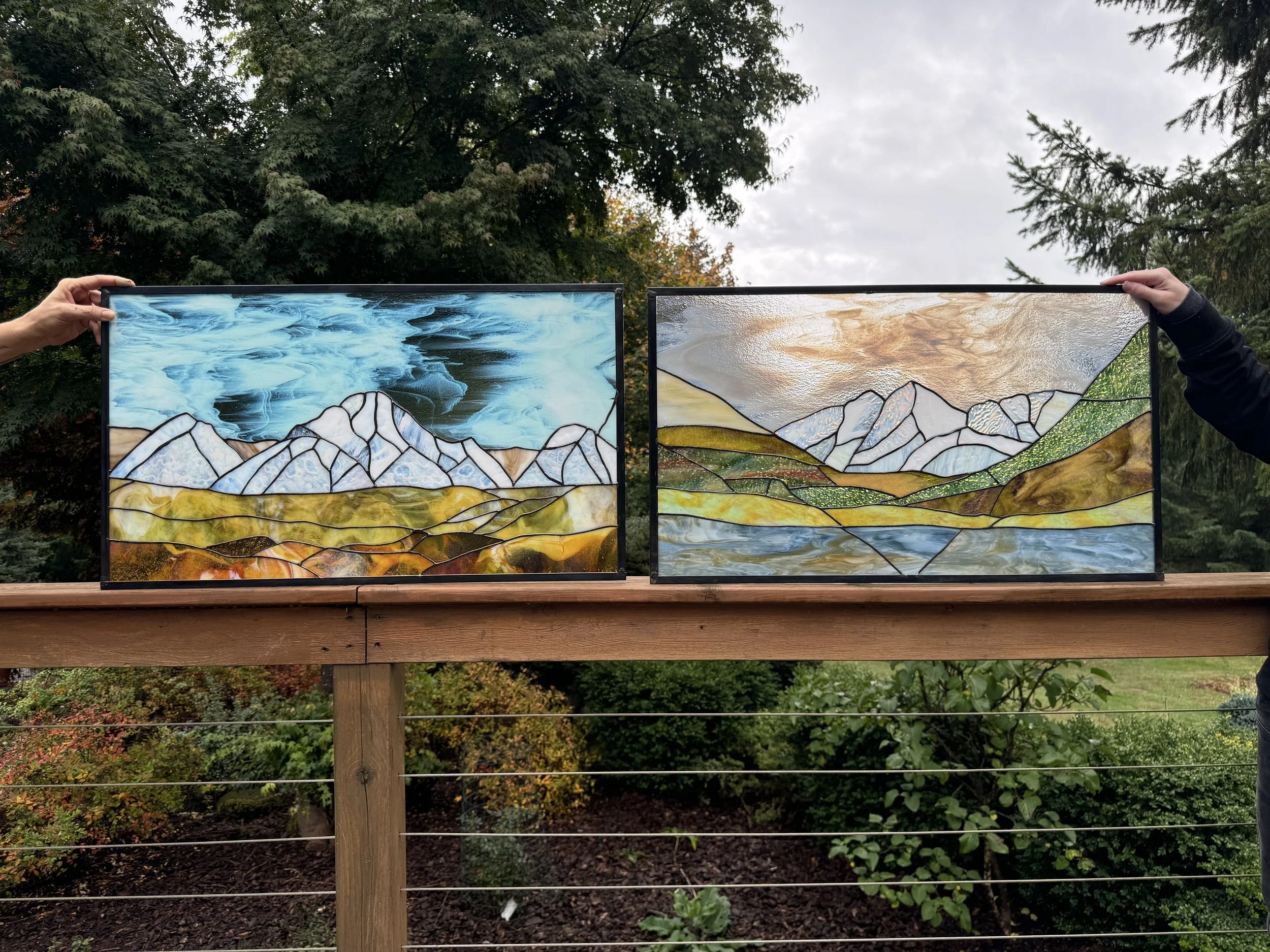 Two stained glass landscape art pieces depicting mountain ranges, clouds, and water, being held up outdoors on a wooden railing.