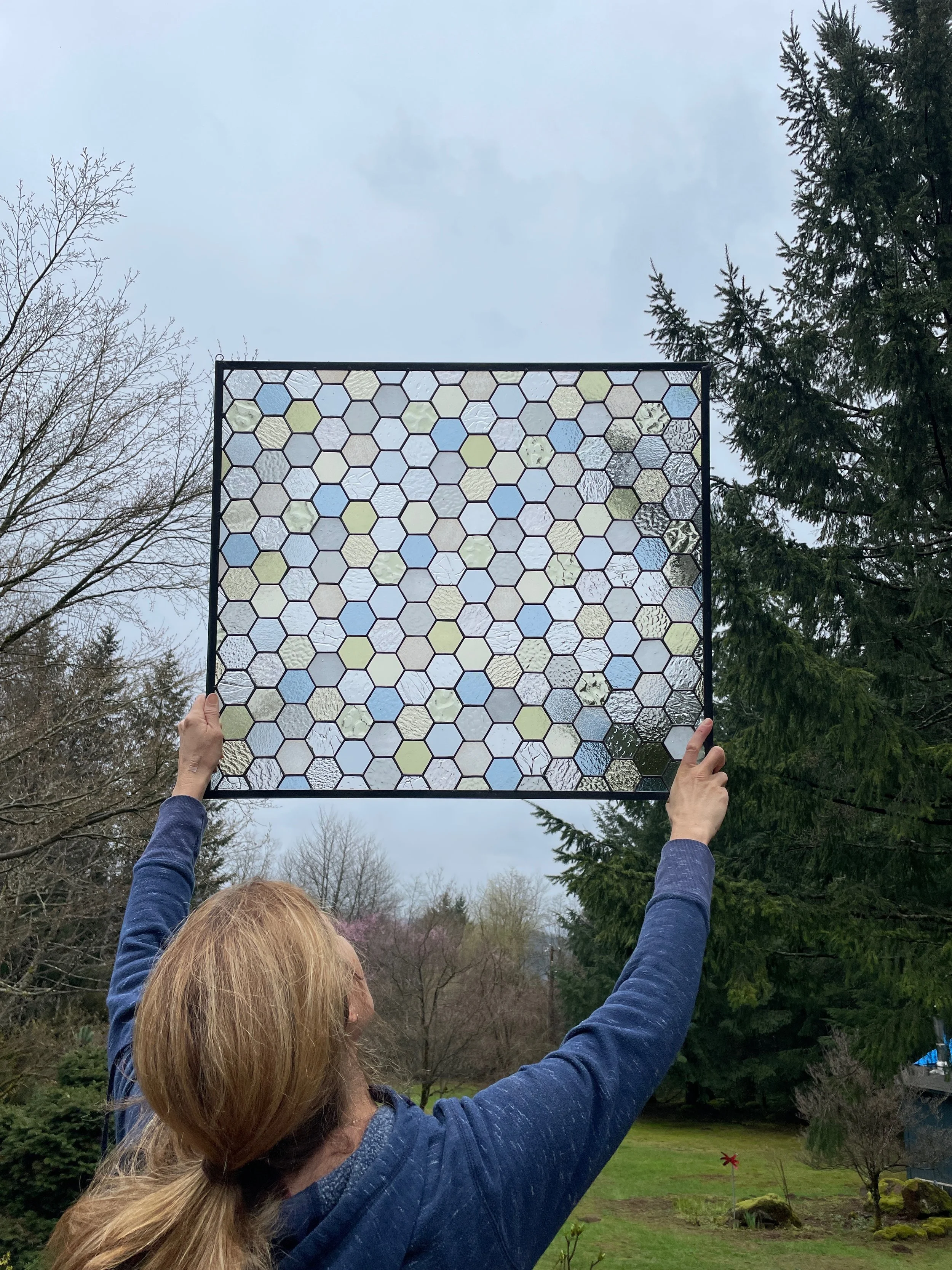 A person holding up a framed rectangular panel with hexagonal glass pieces, with trees and an overcast sky in the background.