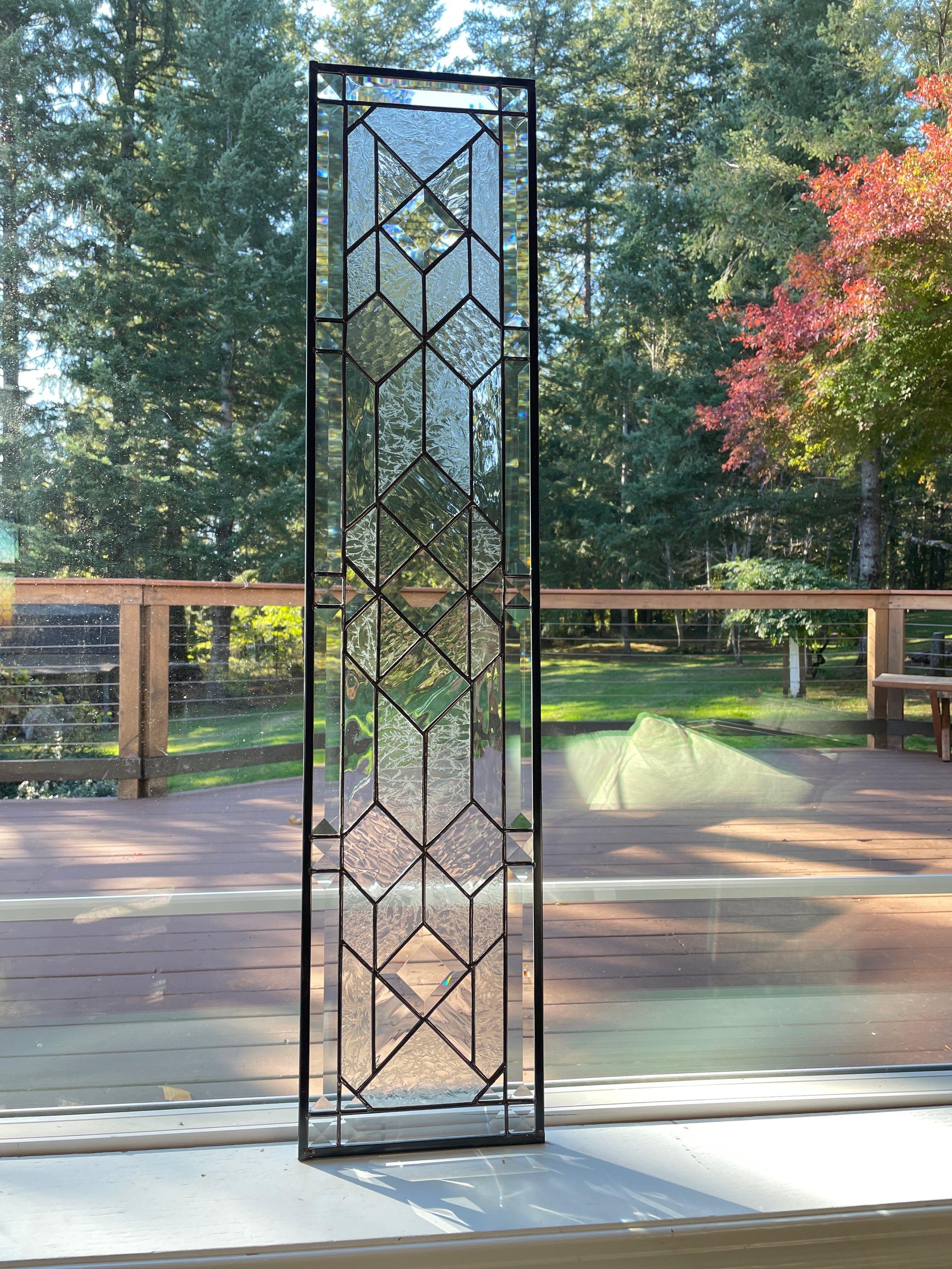 Tall stained glass window with geometric pattern standing on a windowsill. Outside view of a wooden deck, grassy yard, and wooded trees with some red leaves in the background.