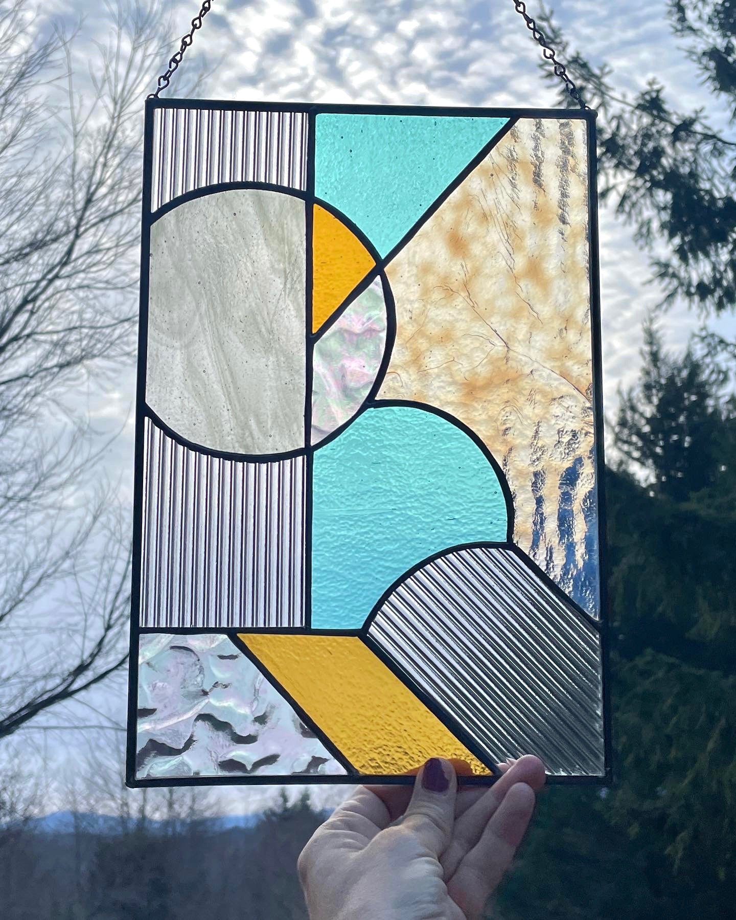 A stained glass panel with geometric shapes in blue, yellow, gray, and black, held up against a cloudy sky with trees in the background.