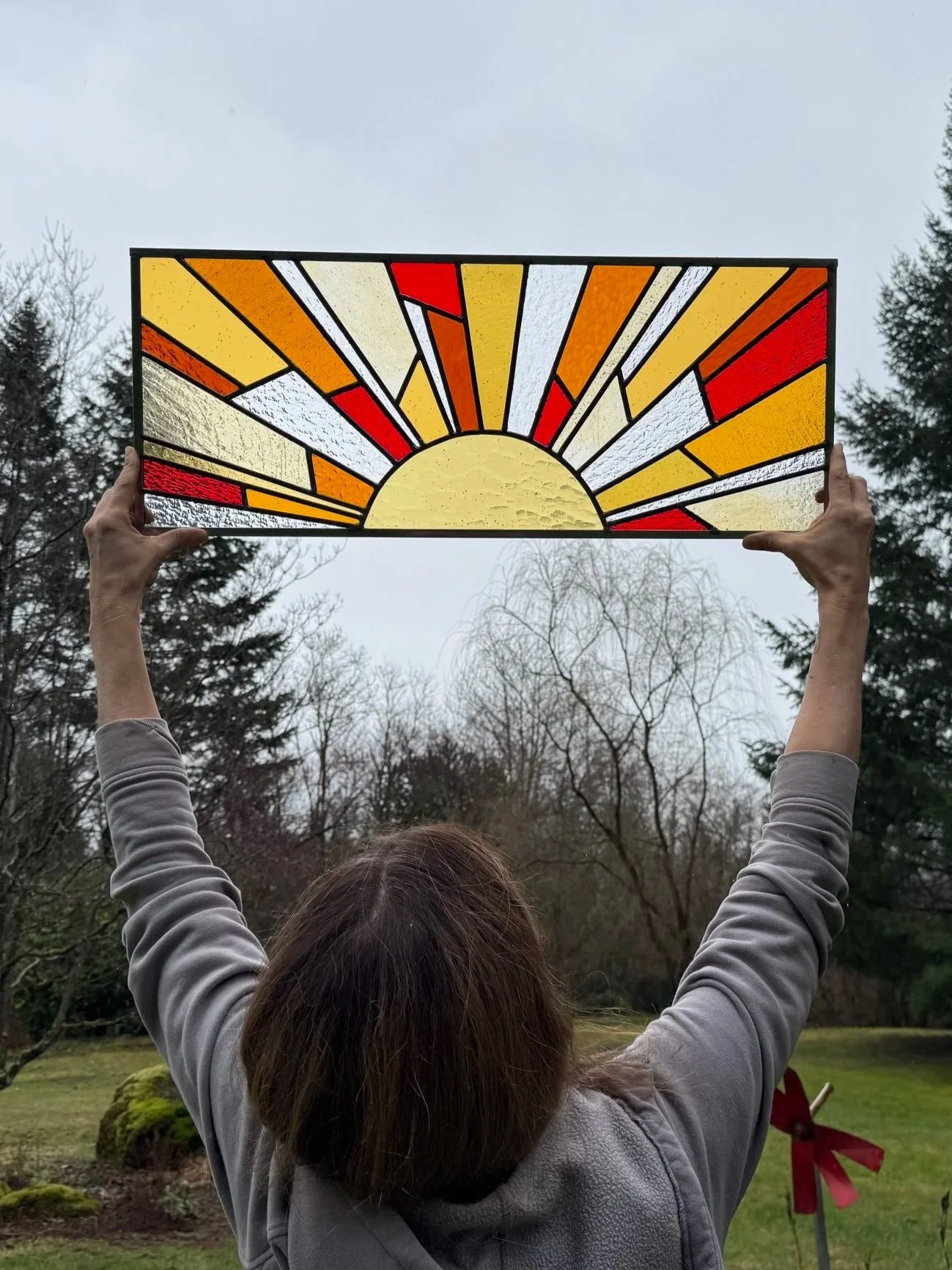 Person holding up a stained glass art piece depicting a sunrise with rays in shades of yellow, orange, red, and white, outdoors with trees in the background.