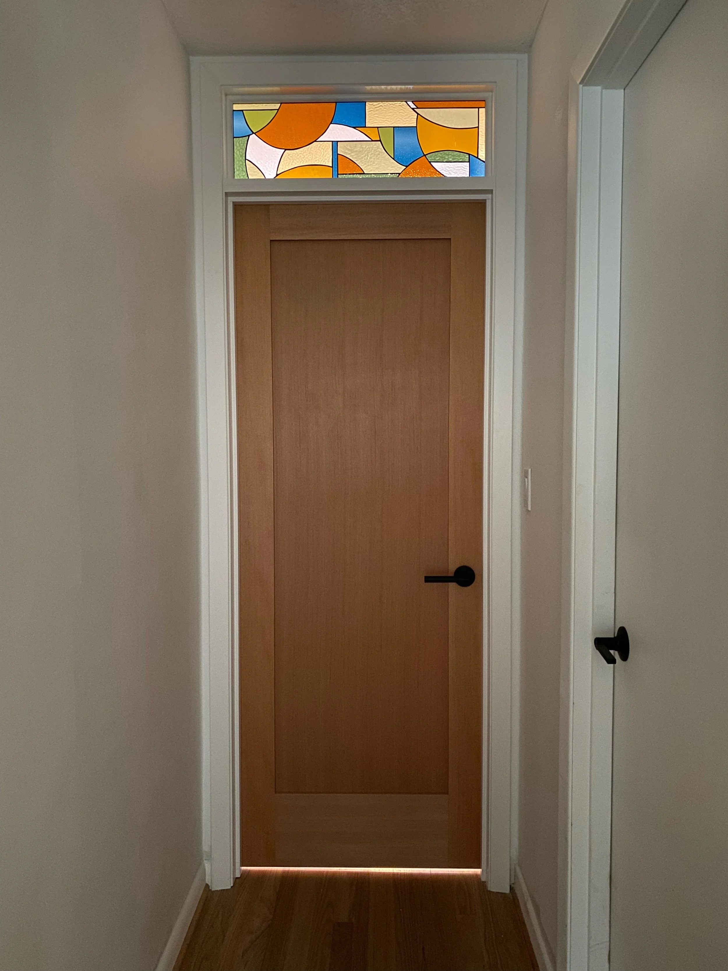 Wooden door with a black handle, surrounded by white trim, with a stained glass window at the top featuring colorful abstract shapes, in a hallway with light-colored walls and wooden flooring.