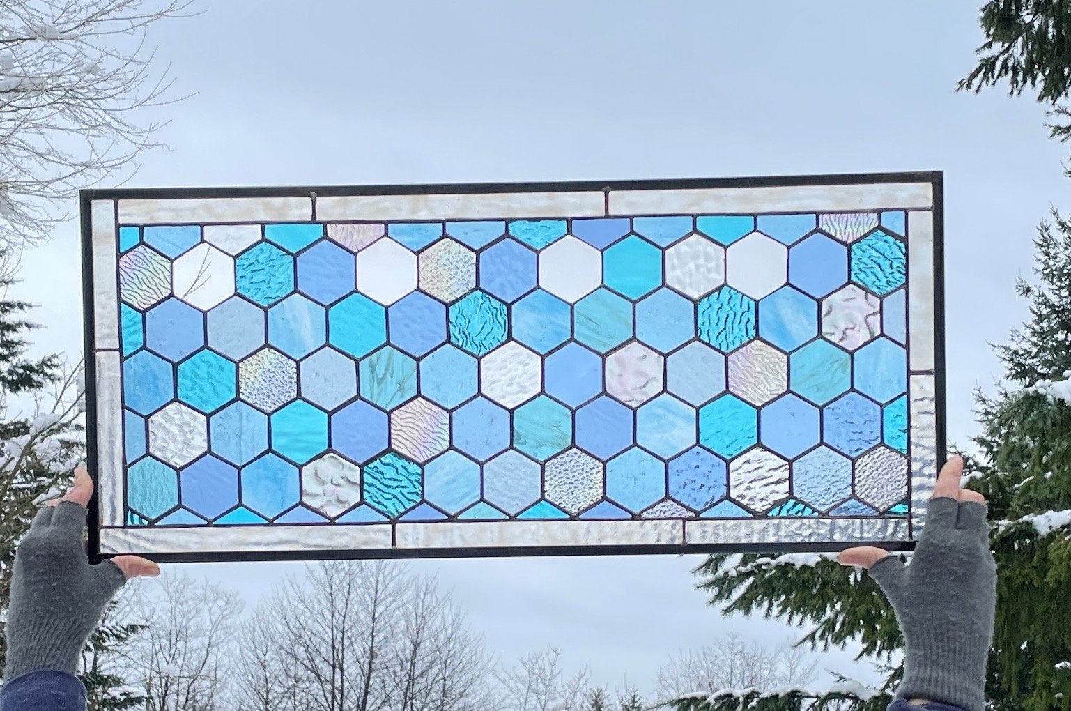 A person holding up a stained glass piece with a honeycomb pattern of hexagonal shapes in various shades of blue, with some patterned and textured glass pieces, against a cloudy sky with trees in the background.