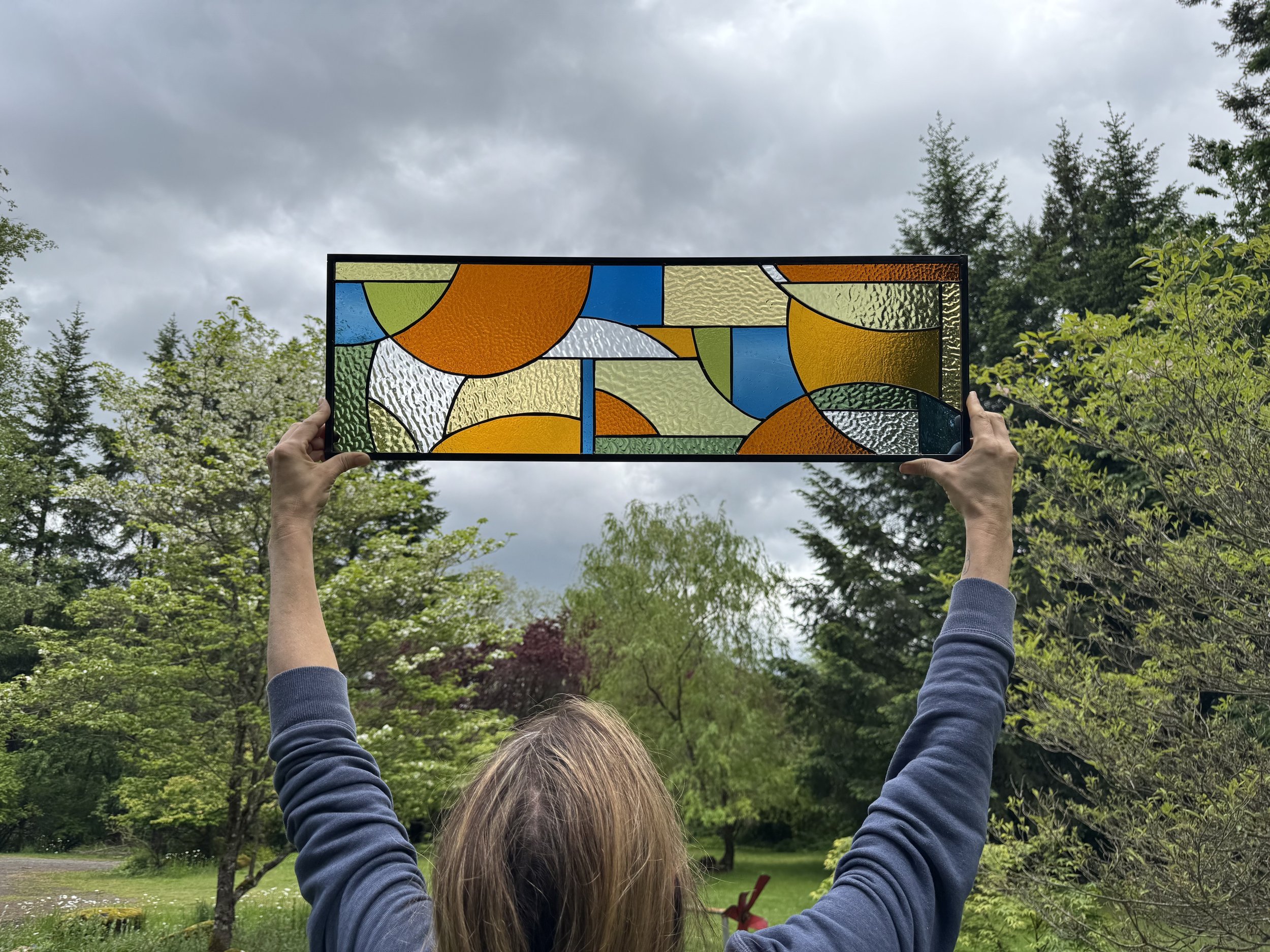 Person holding up a colorful abstract stained glass panel outdoors in a green park with trees and cloudy sky.