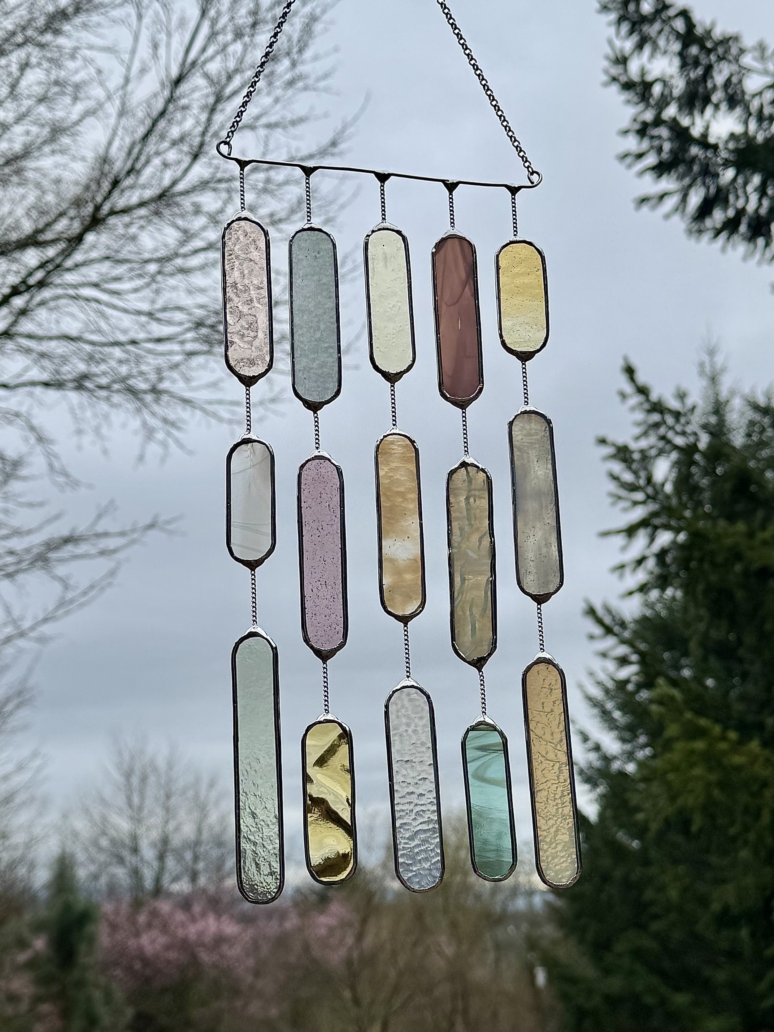Colorful stained glass wind chime hanging outdoors with trees and cloudy sky in the background.