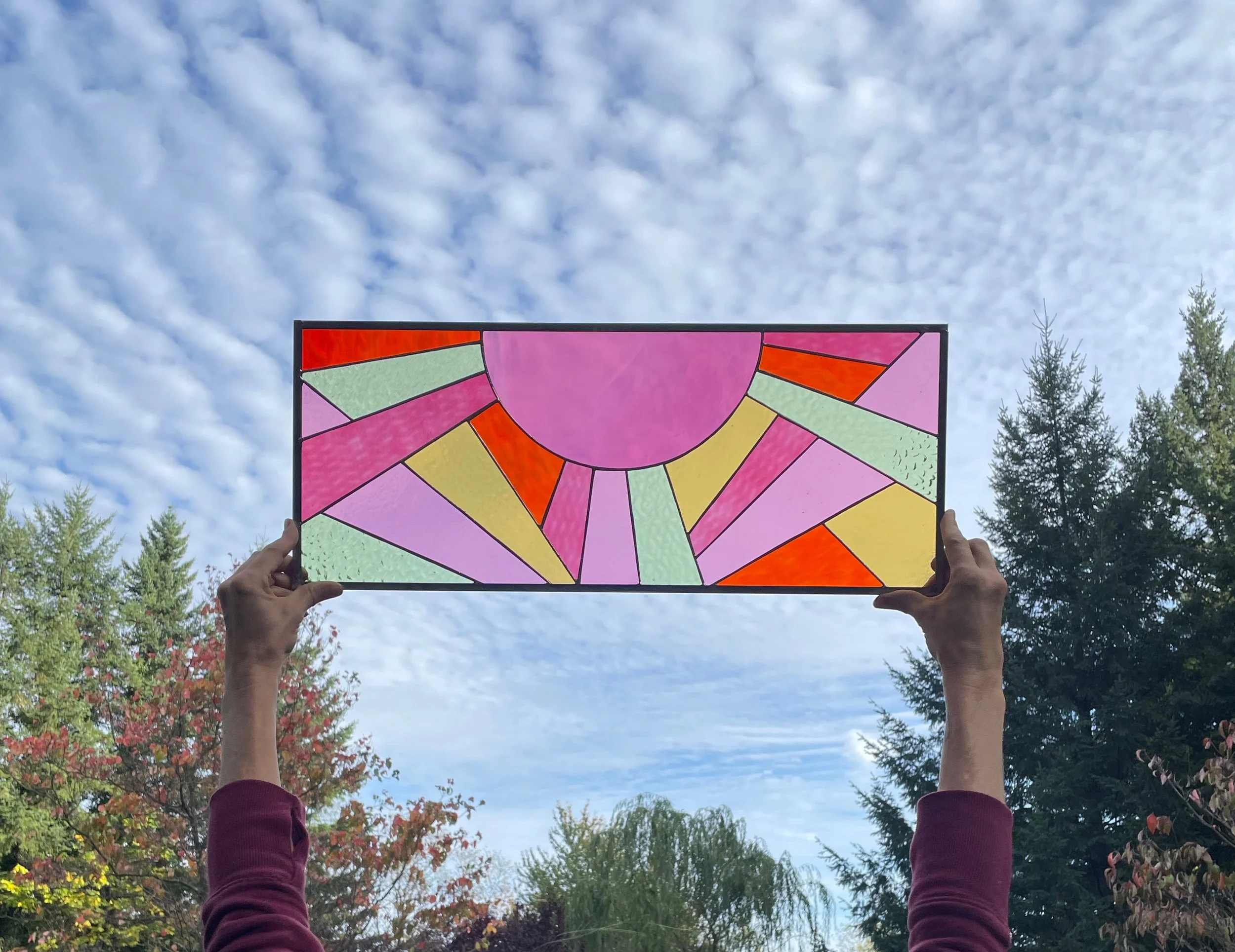 A person holding a colorful stained glass window panel up to the sky, with trees and a partly cloudy sky in the background.