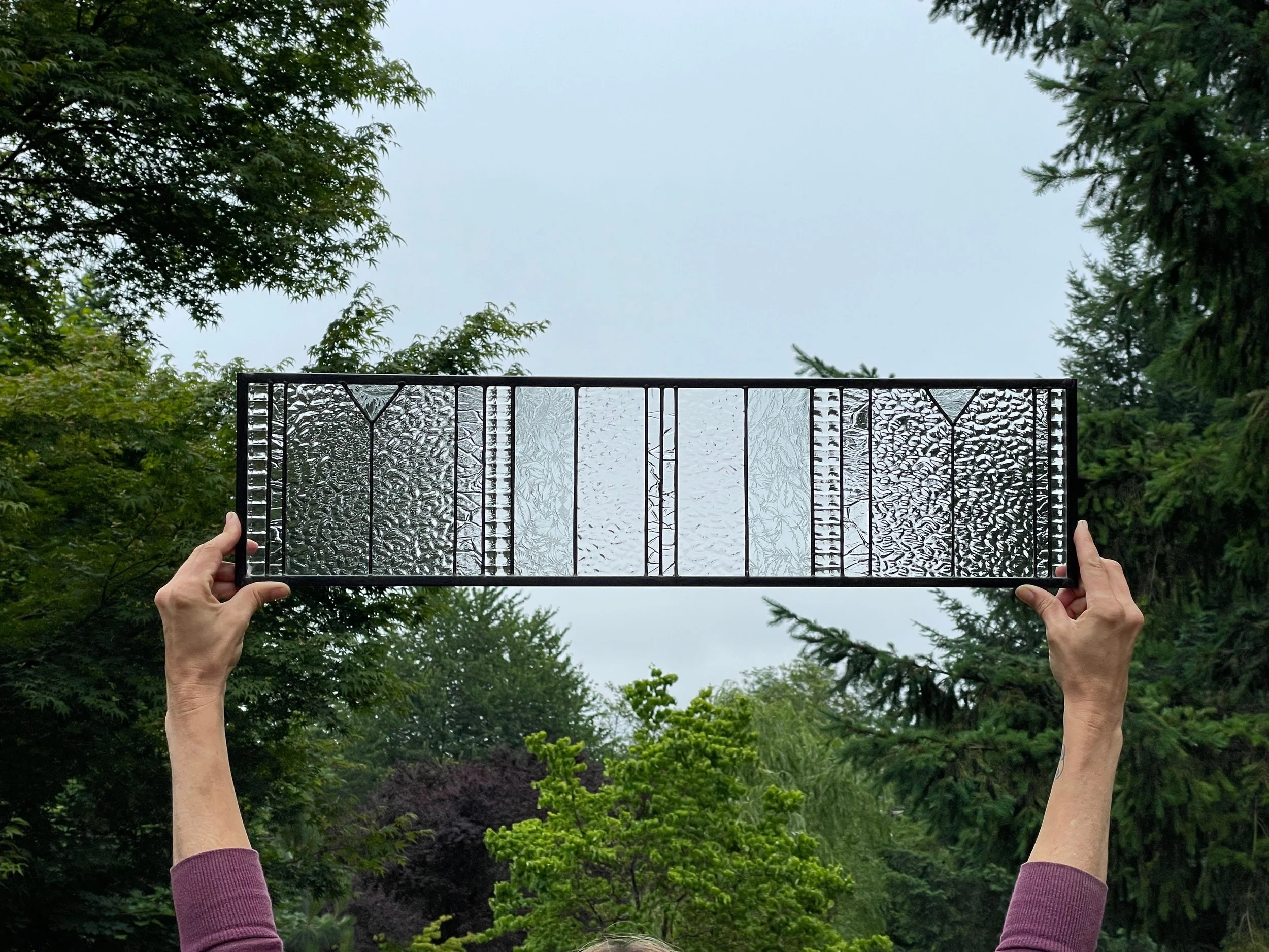 Person holding up a large textured glass window outdoors with trees and an overcast sky in the background.