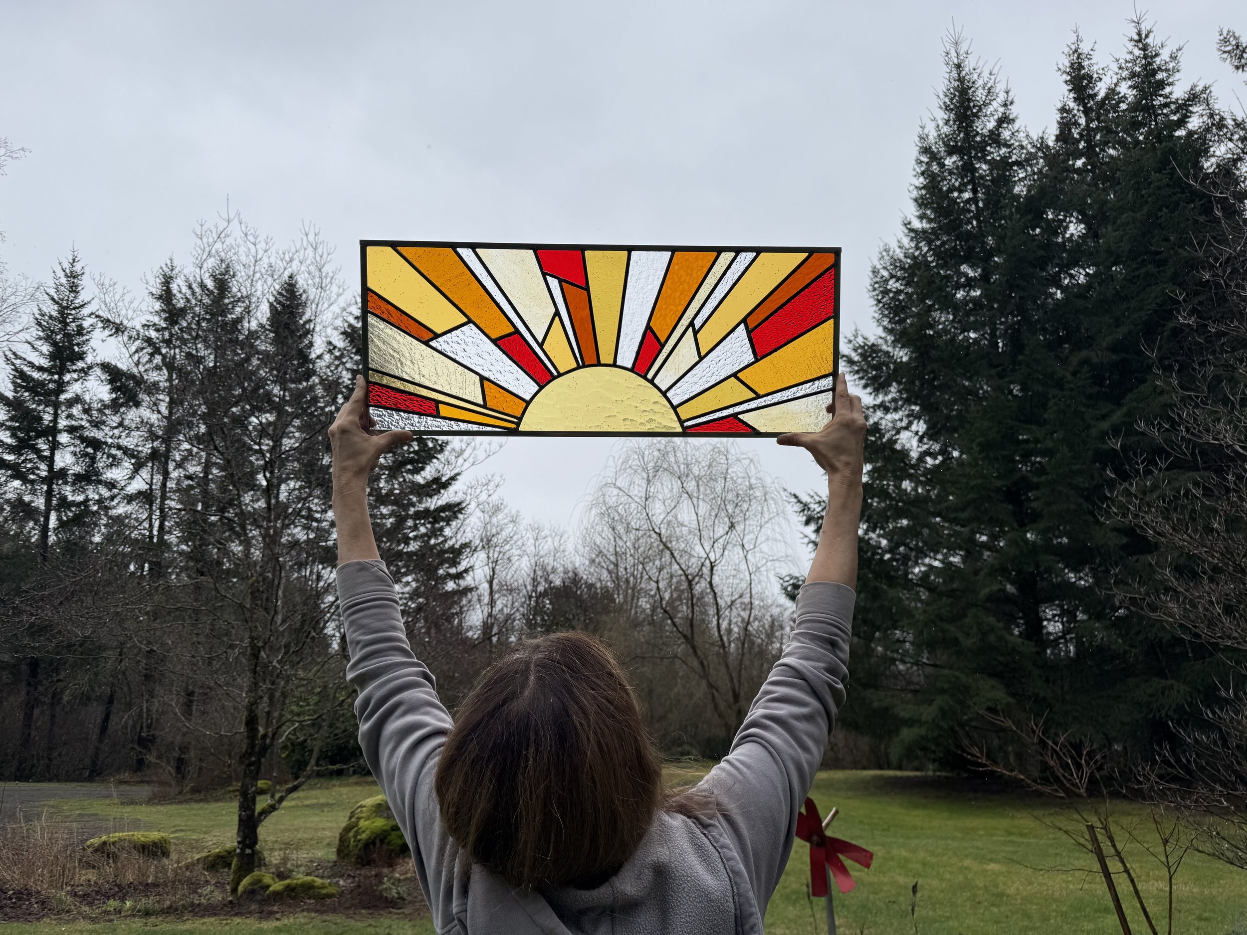 Person holding up a stained glass panel depicting a sunrise with rays of sun in orange, yellow, and white against an outdoor background with trees and grass.